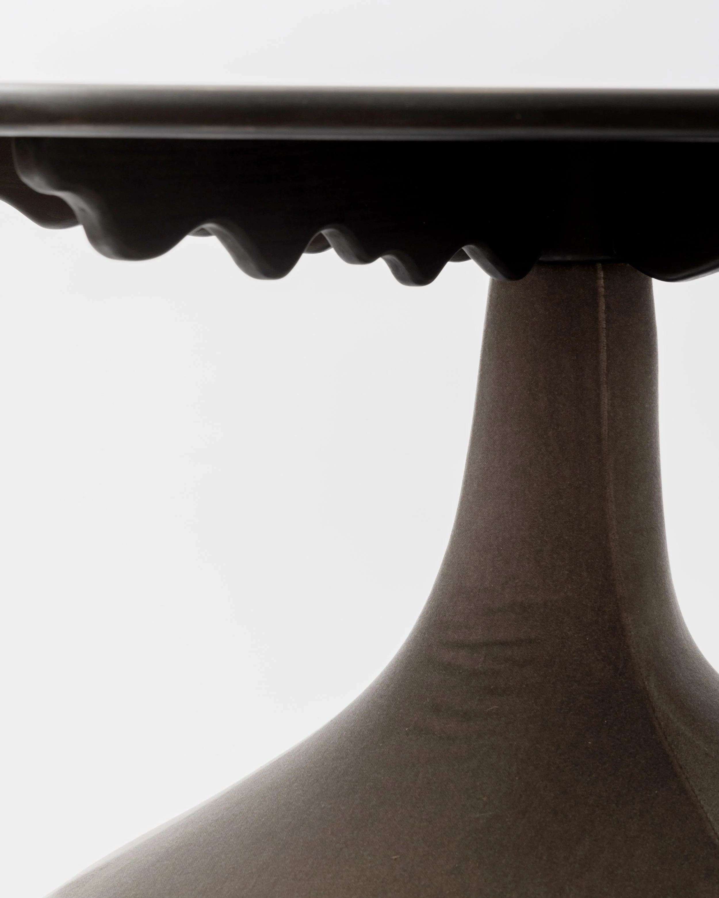 Close-up of a dark brown or black ceramic or pottery piece with ridged and curved design, set against a plain light gray background.