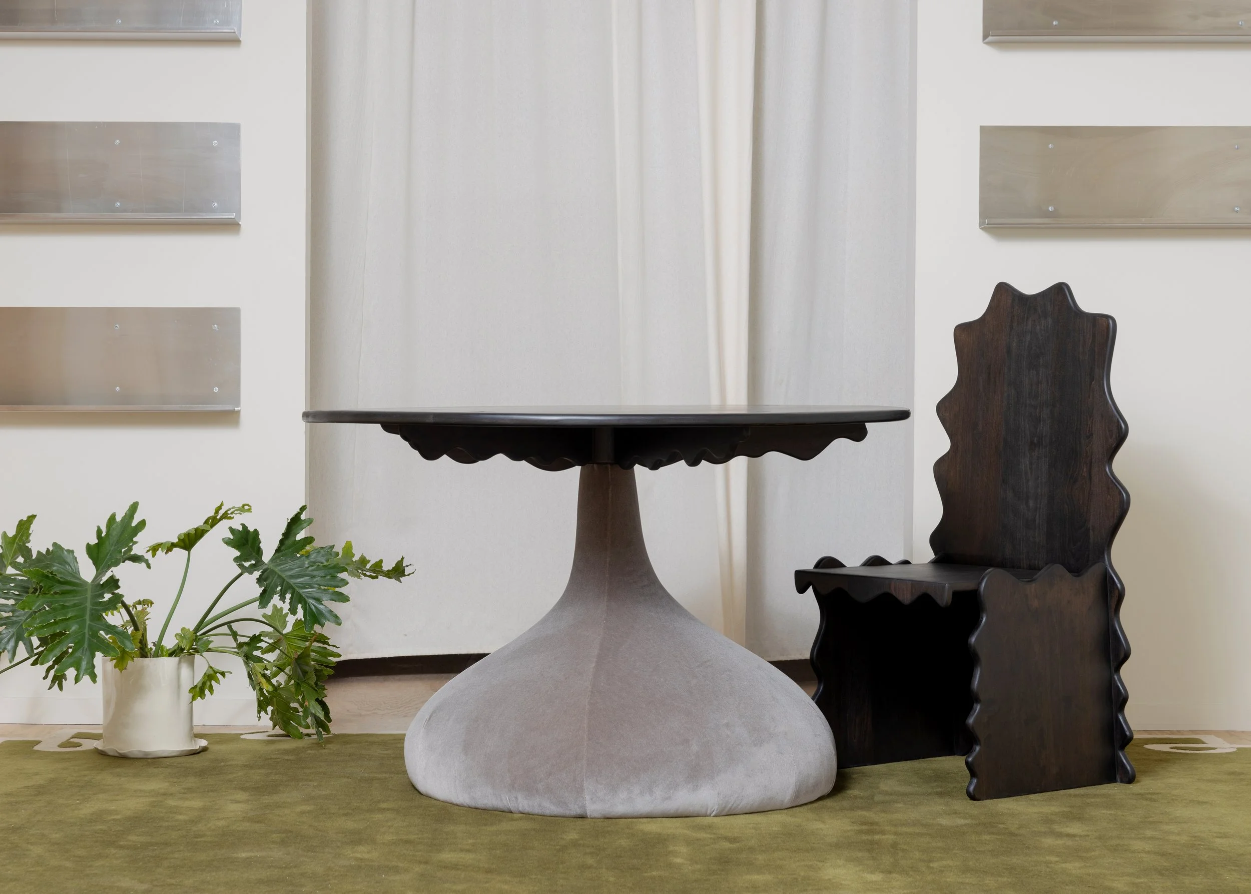 A black wooden chair with a wavy backrest visible beside a table with a rounded, scalloped edge. A potted green plant with large leaves is positioned on the floor nearby. The background features four rectangular metal nameplate holders on a light-col