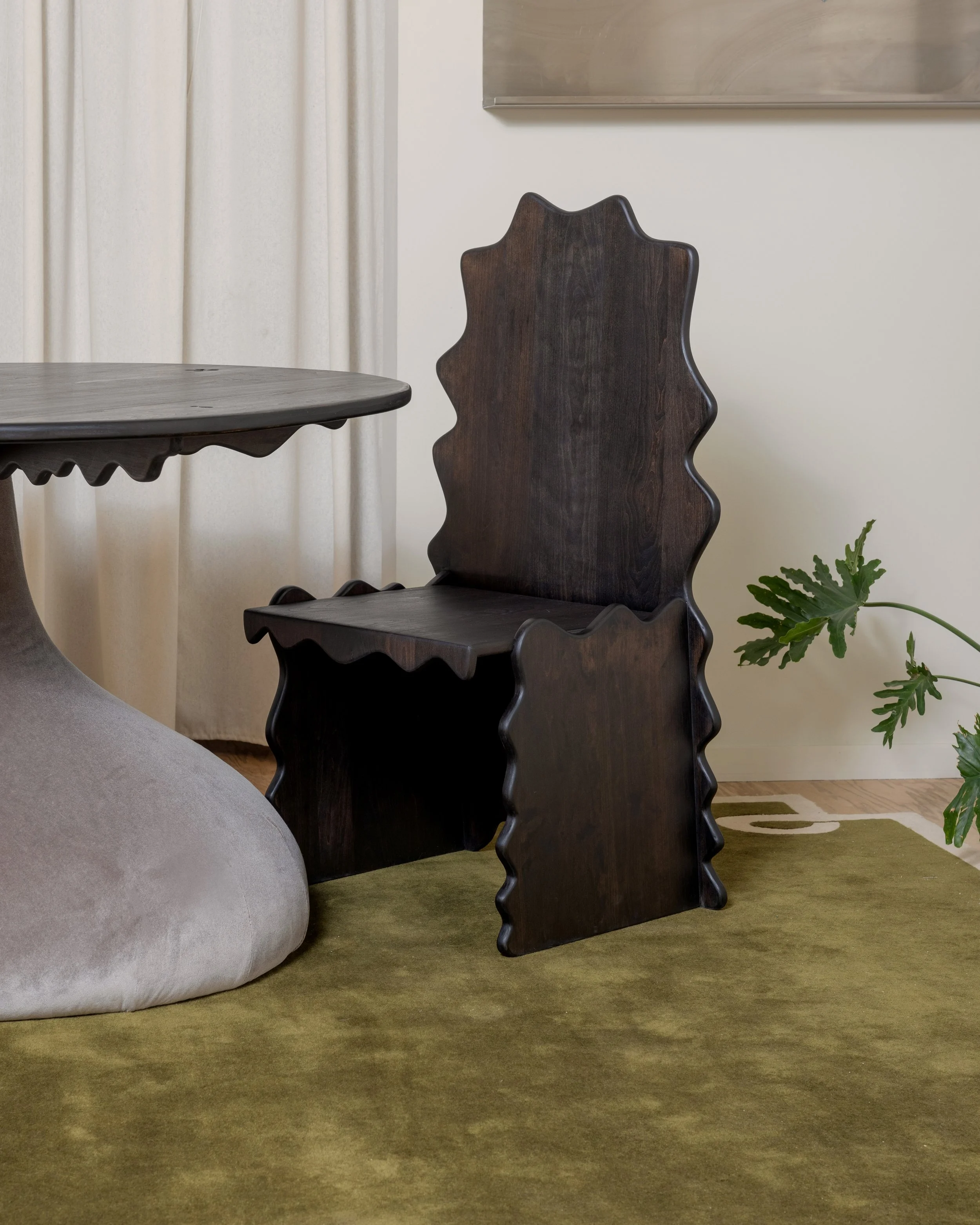 Close-up of a wooden chair with a wavy backrest, partially behind a light gray sofa and a table, in a room with a green carpet and a plant in the corner.