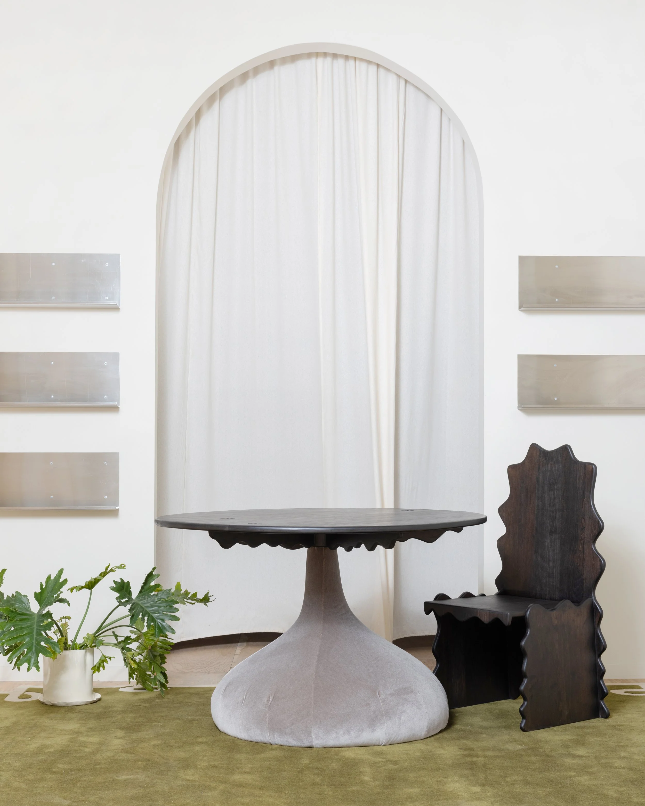 Interior room with white walls and a large arched window covered by white curtains. There is a round table with a pedestal base, a black ornate wooden chair to the right, and a potted green plant on the left on a green rug.