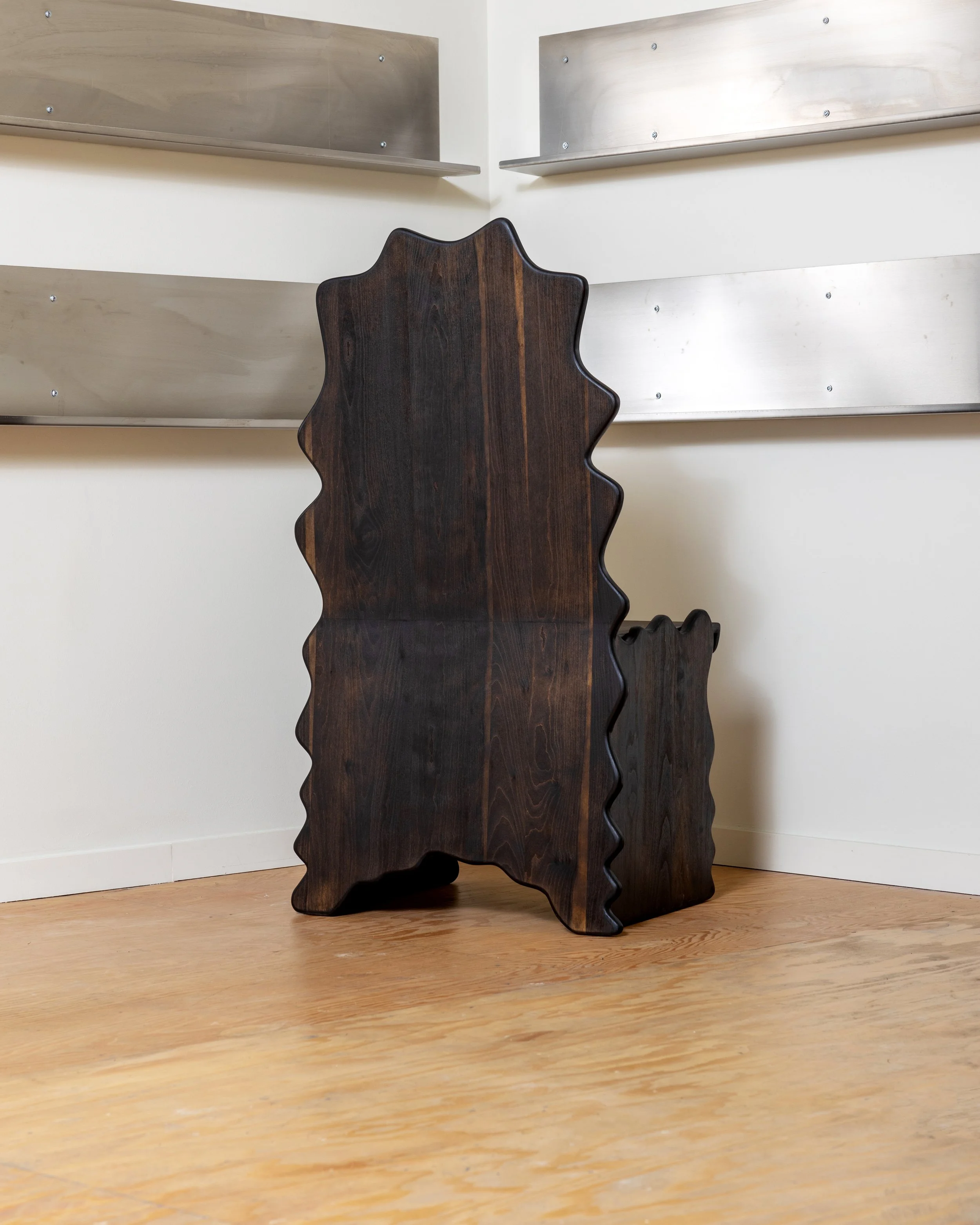 A wooden dresser with a unique zigzag edge design is situated on a wooden floor next to a white wall. Behind it, there are two metal shelves attached to the wall, one directly above the other.