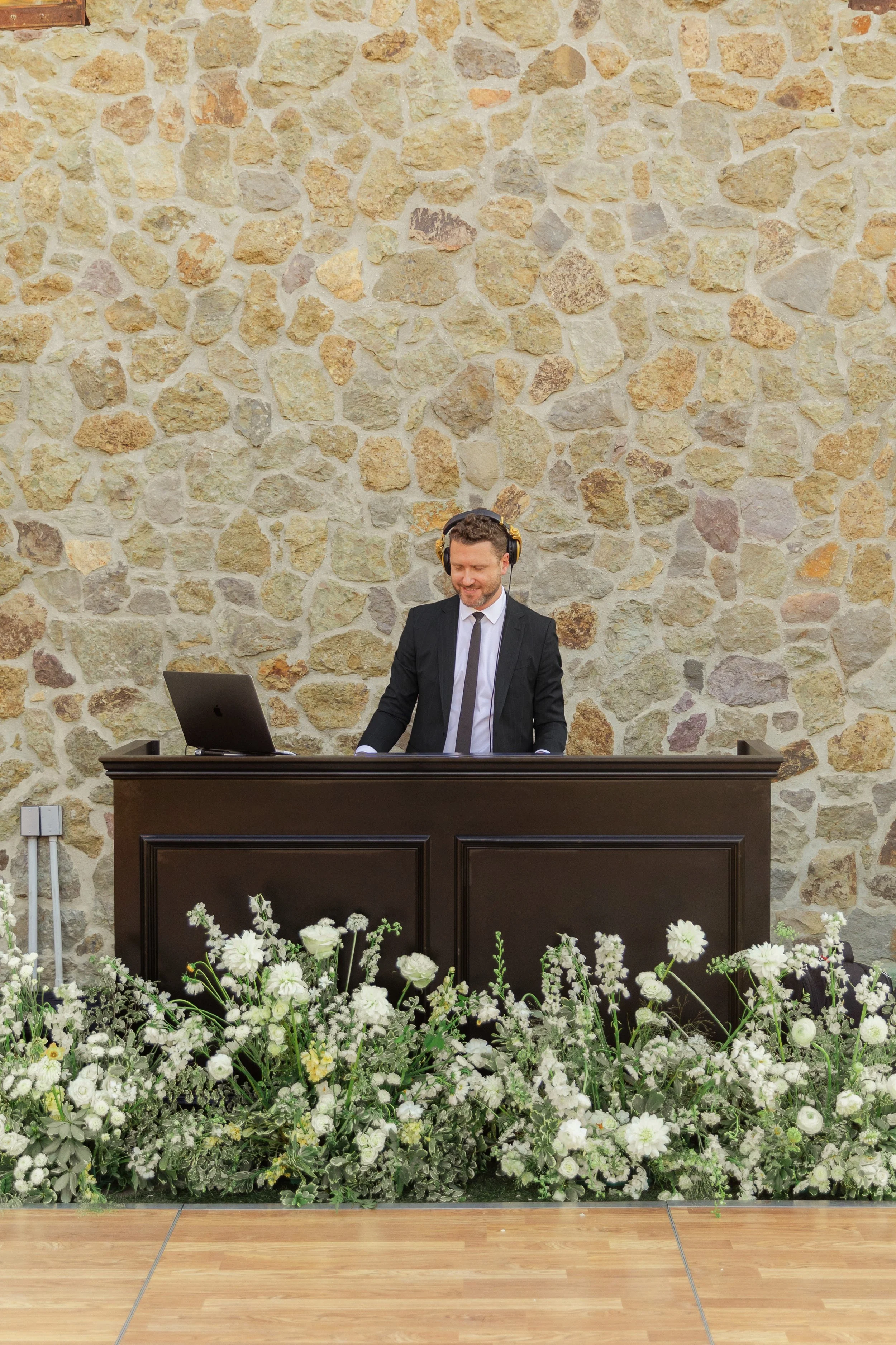 A man in a black suit and tie, wearing headphones, standing behind a dark wooden DJ booth, smiling, with white flowers and greenery in front of the booth, against a stone wall background.