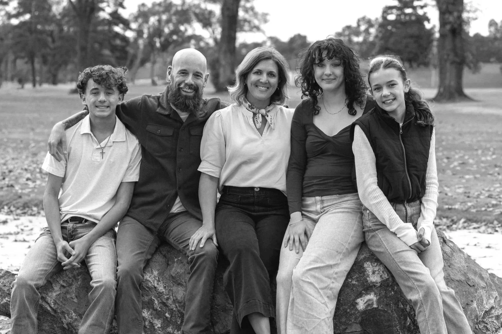 A black and white photo of a family of five sitting on a large rock in a park with trees in the background, smiling at the camera.