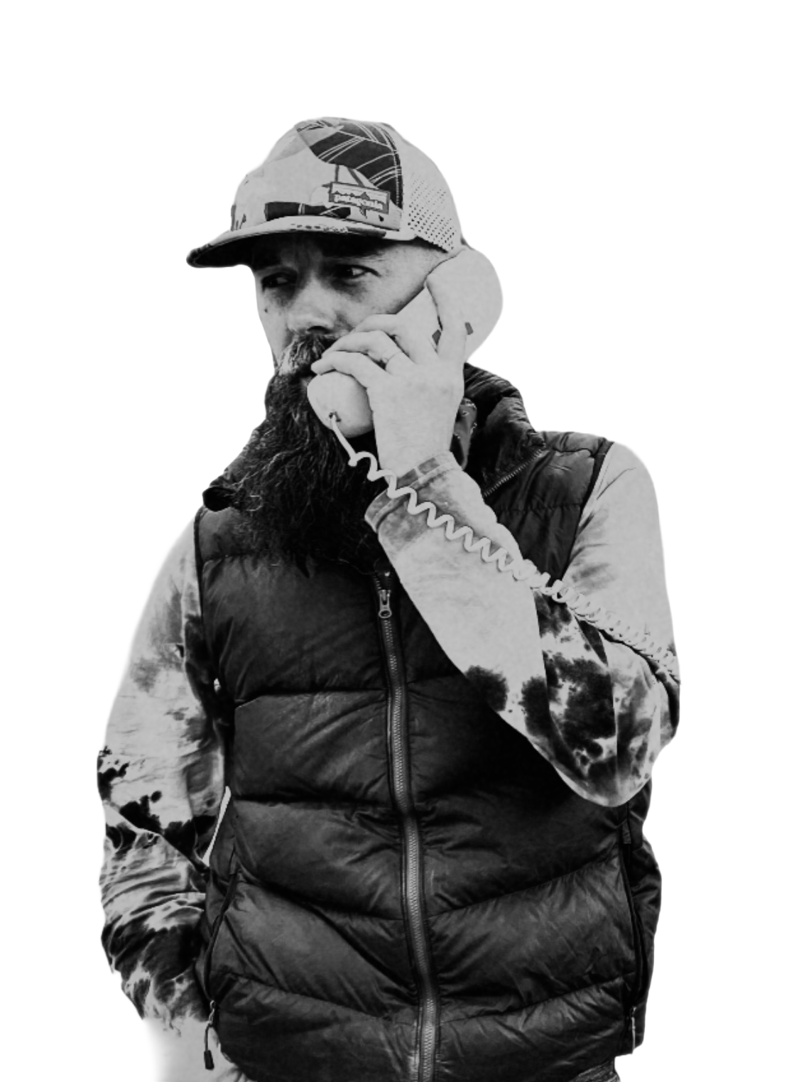 Black and white photo of a man with a beard and mustache speaking on a landline phone, wearing a baseball cap and a puffy vest with camouflage sleeves.