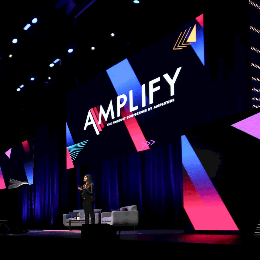 A woman speaking on stage at a conference called Amplify with the subtitle "The Product Conference by Amplify" displayed on large screen behind her, with chairs and a table on stage.