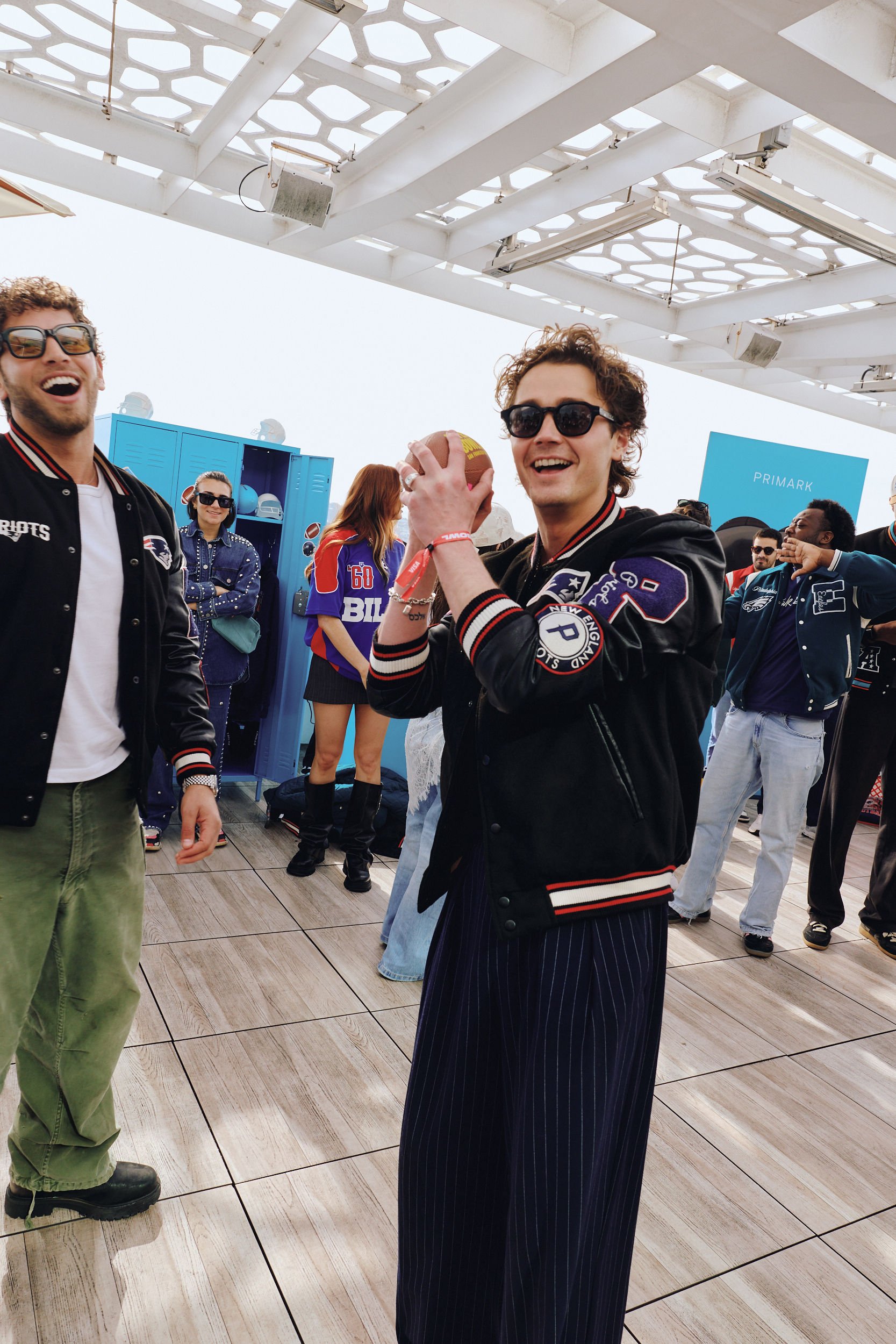 A group of young people at a rooftop gathering, with a man in a black varsity jacket holding a football, smiling and wearing sunglasses. Others in the background are dressed casually, some wearing sports jerseys and sunglasses, with blue lockers and a partially covered sky overhead.