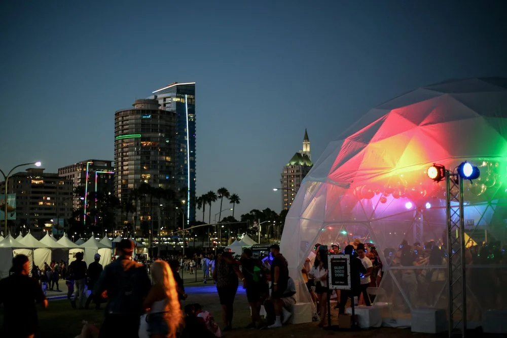 Night scene of an outdoor event with a geodesic dome stage illuminated by colorful lights, people gathered around, and city buildings with lights in the background.