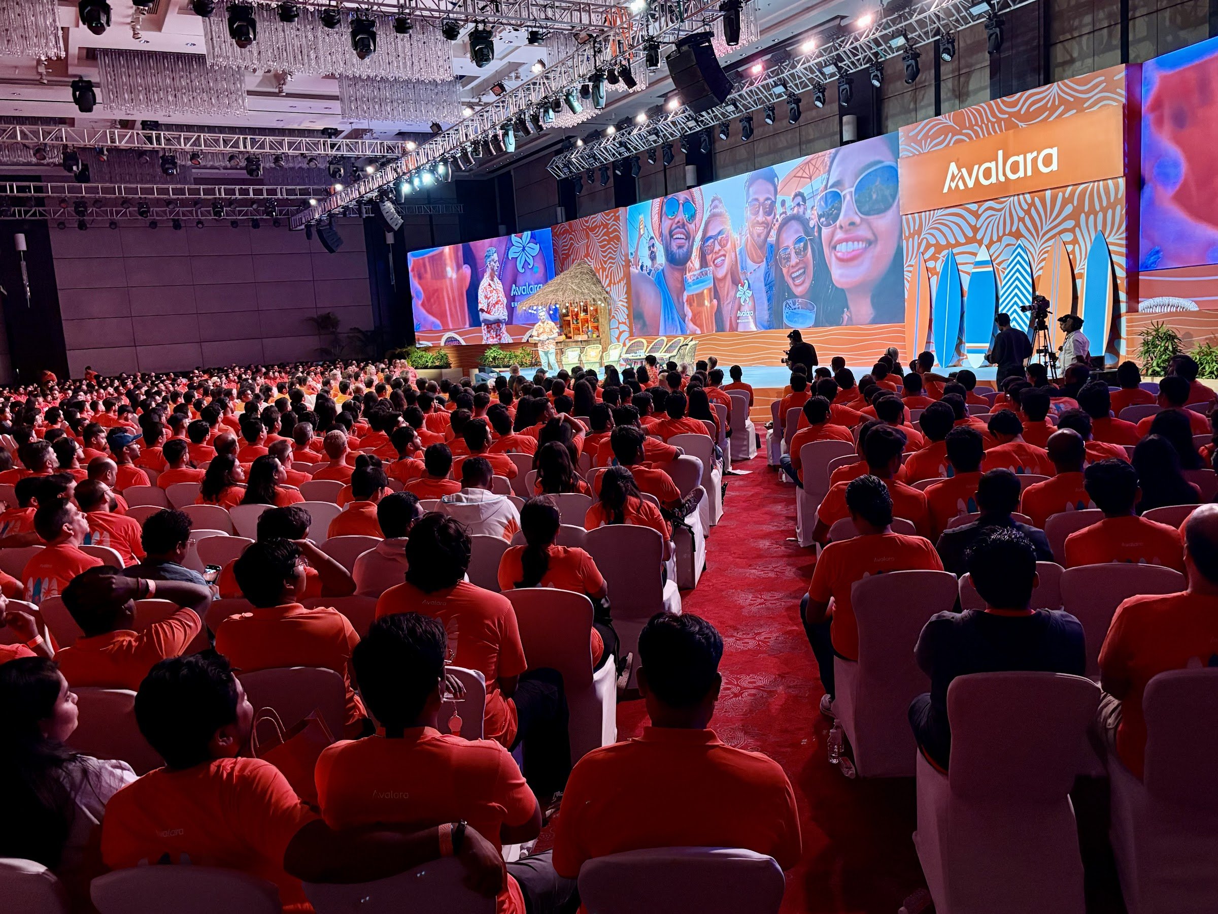 A large conference room filled with people wearing red shirts, facing a stage with a large screen displaying the Avalon logo and images of smiling people at a tropical-themed event.