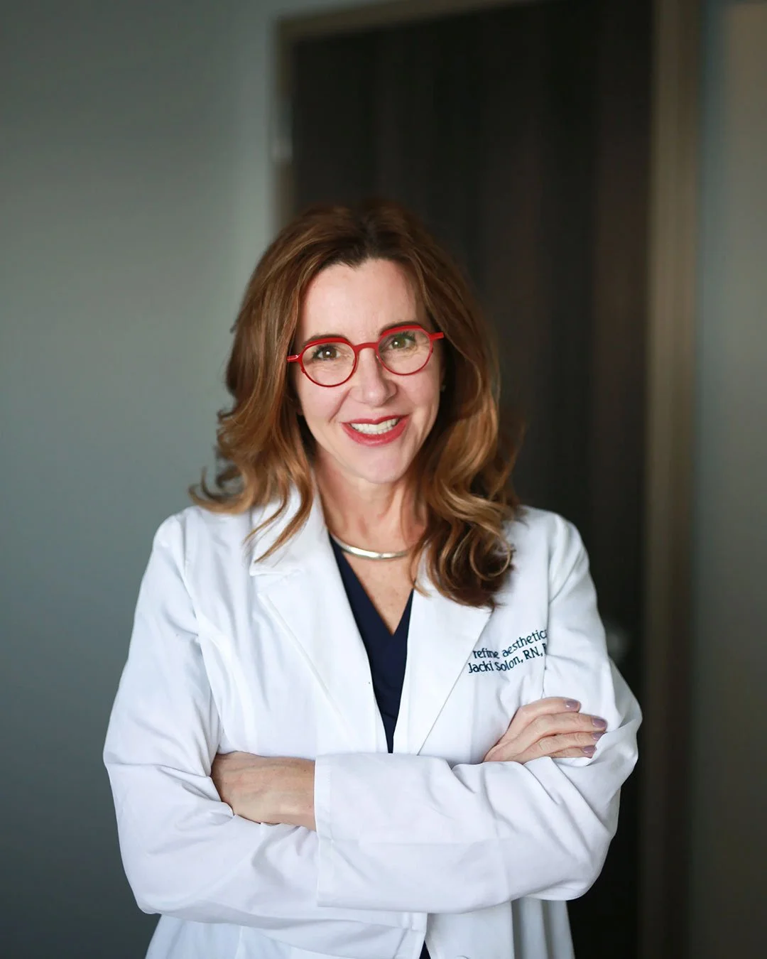 A smiling woman with red hair, red glasses, and light skin posing with crossed arms in a white lab coat in an indoor setting.