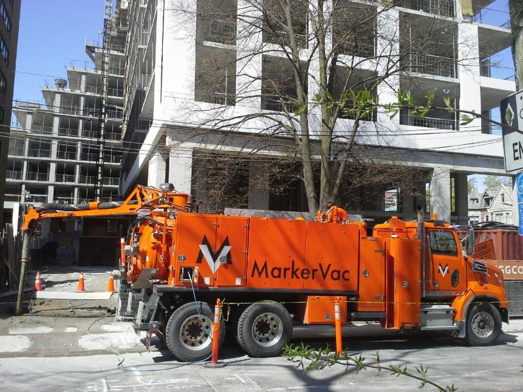 Orange MarkerVac vacuum truck parked at a construction site with a partly constructed building in the background.