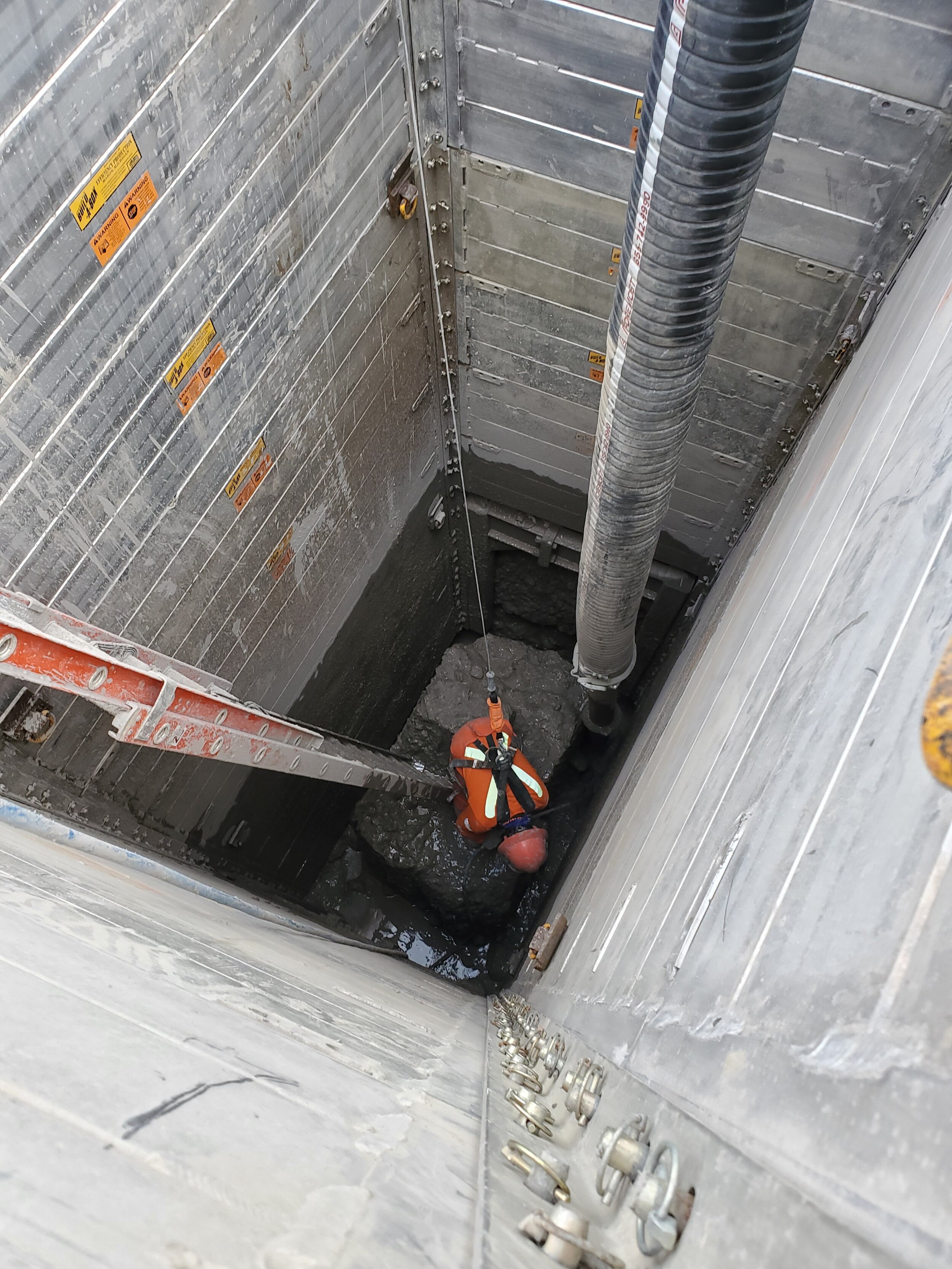 A person wearing a safety harness and hard hat is working inside a deep, narrow construction pit with metal walls, utilizing a ladder and large hose for support.