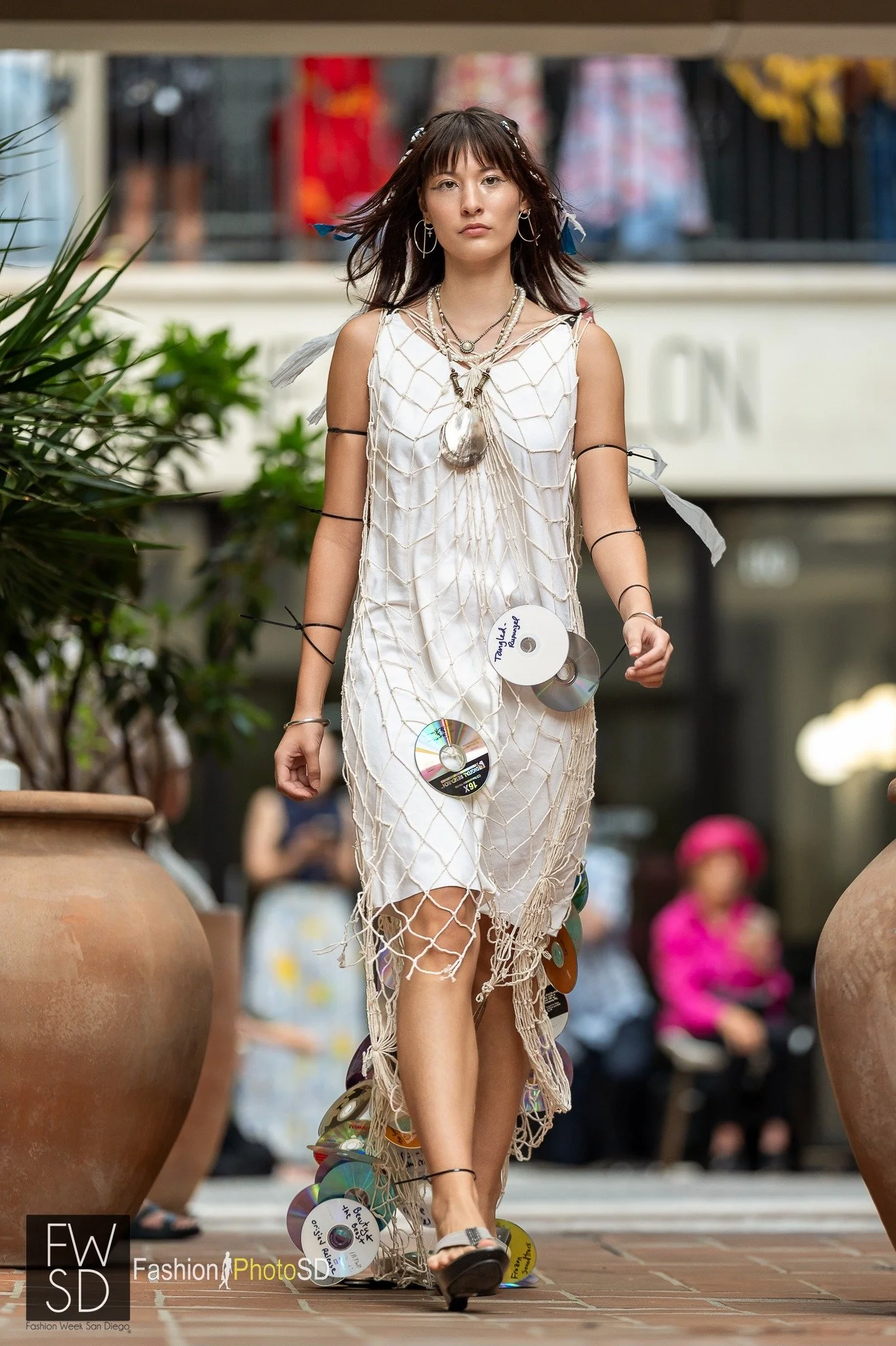 A young woman walking on a runway during a fashion show, wearing an unconventional dress made of CDs and a net-like fabric. She has layered necklaces and earrings, with loose hair and a serious expression.