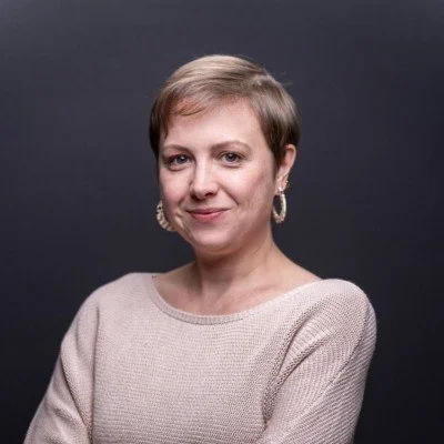 A portrait of Ellen, a white woman with short blonde pixie. She has a closed smile and is wearing golden hoops alongside a beige top, set against a charcoal grey backdrop.
