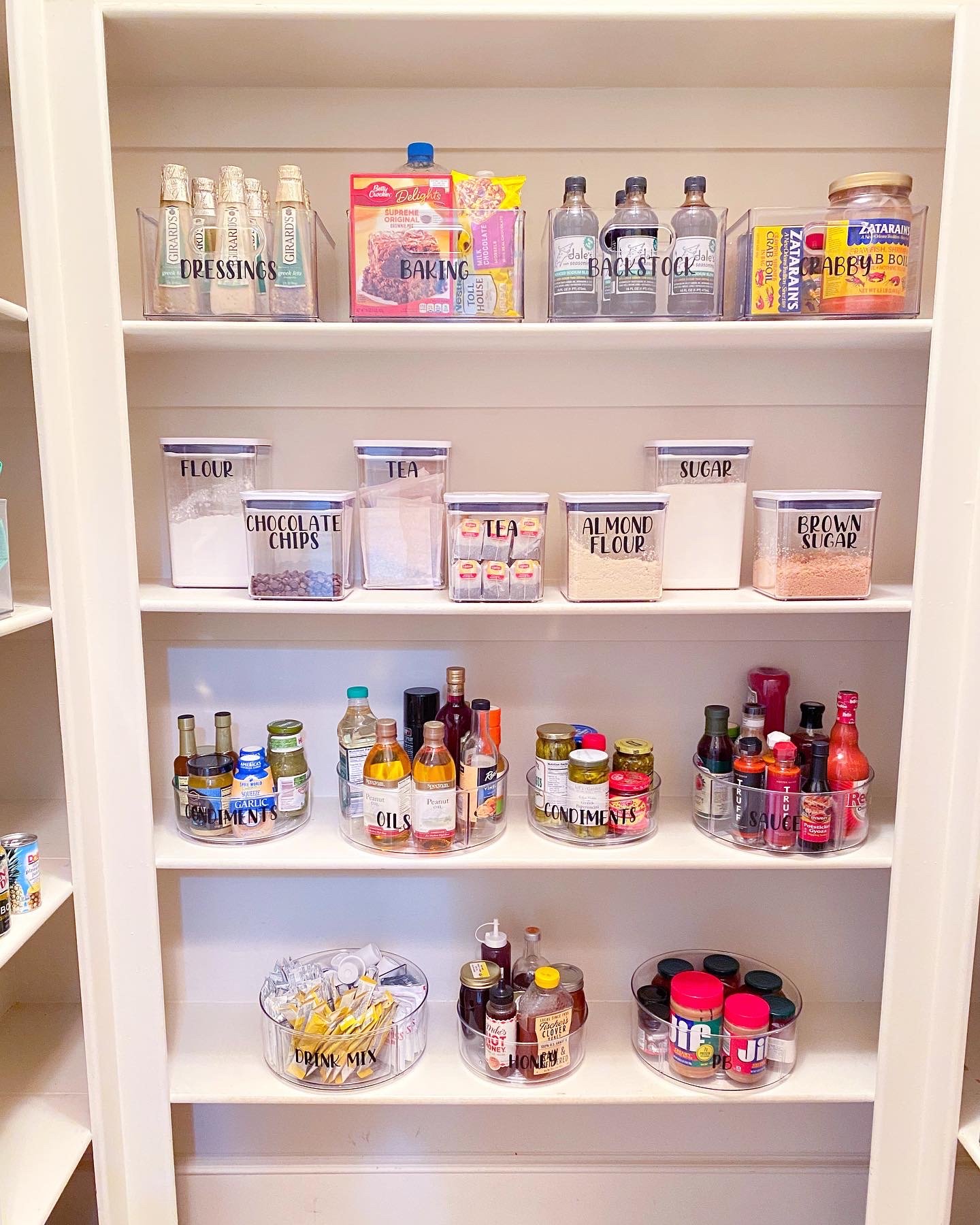 A pantry shelf organized with labeled containers and bottles for baking ingredients, dressings, stock, candy, oils, condiments, drink mix, and honey.