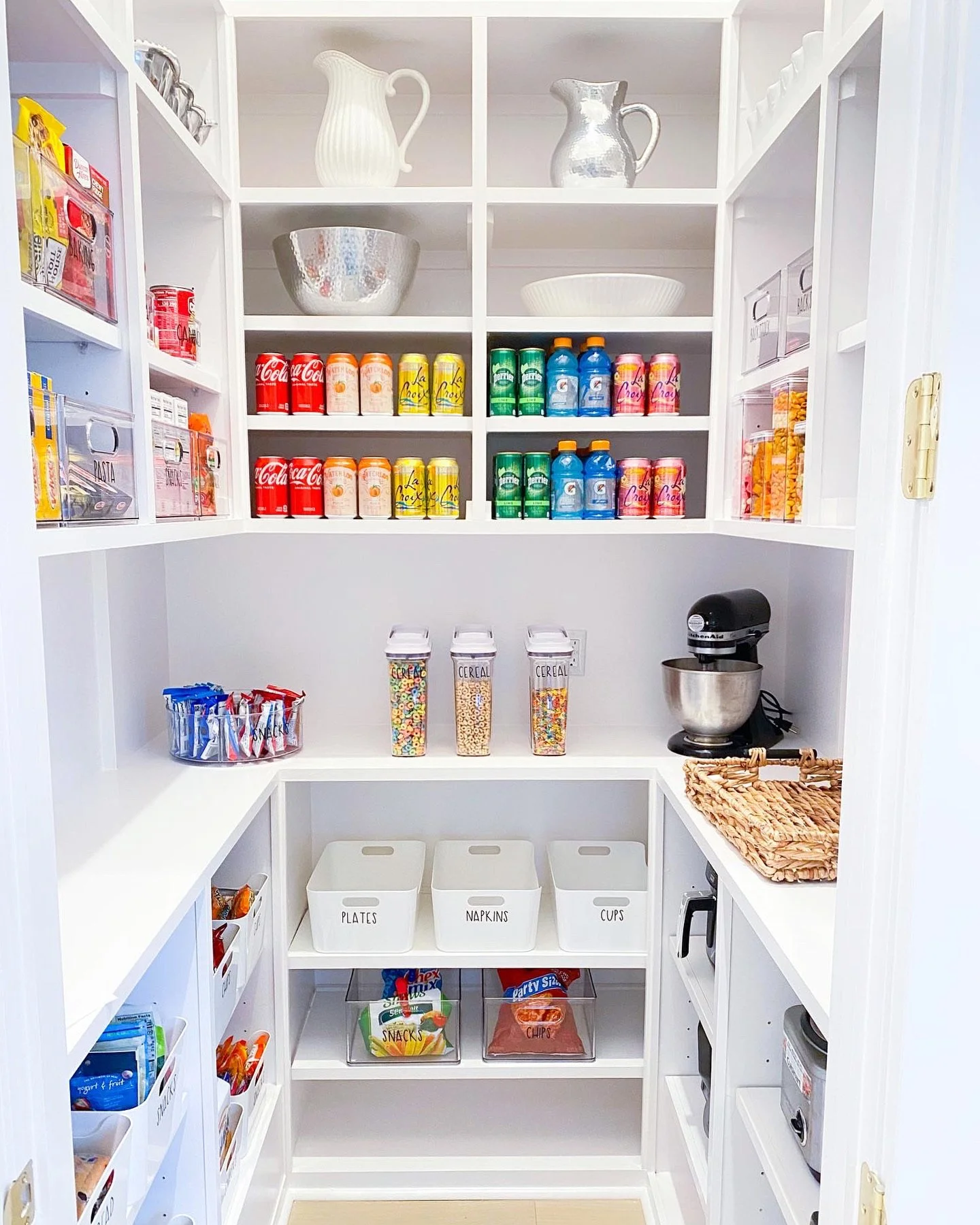 Organized white pantry with shelves holding canned drinks, cereal boxes, snacks, baking ingredients, a stand mixer, bowls, and labeled storage bins for plates, napkins, and cups.