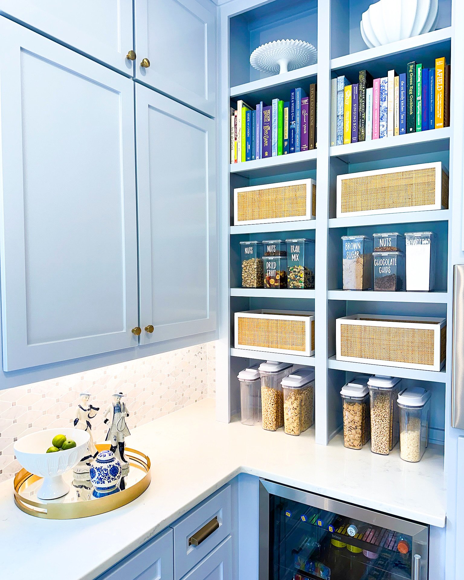 Kitchen pantry with light blue cabinets, shelves filled with books, labeled jar containers of nuts, dried fruit, trail mix, brown sugar, chocolate chips, and pasta, white countertop with a bowl of limes, decorative figurines, and an adjoining wine fridge.