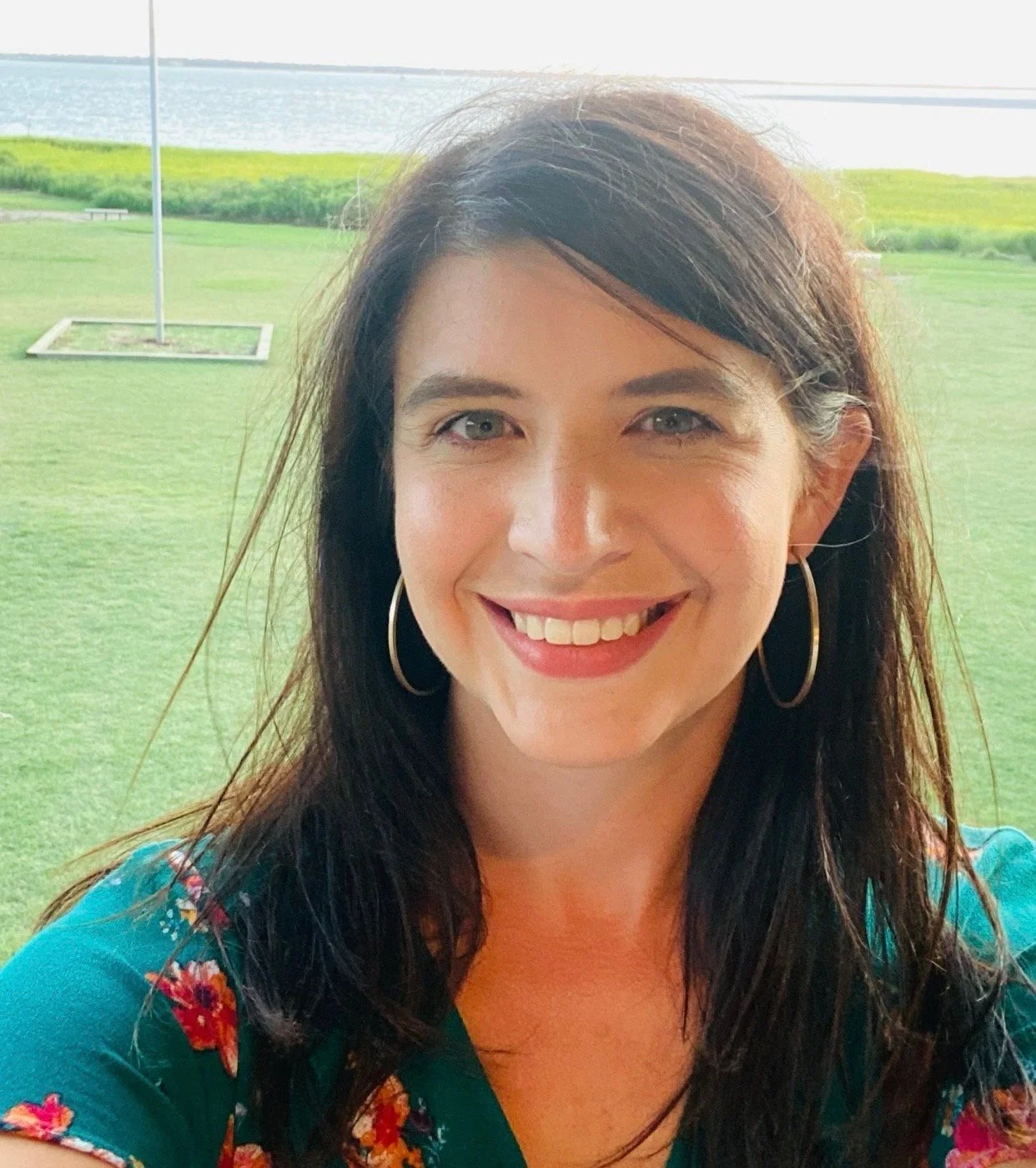 A woman with shoulder-length dark hair, wearing a teal dress with a floral pattern and hoop earrings, smiling outdoors with grassy area, a flagpole, and water in the background.