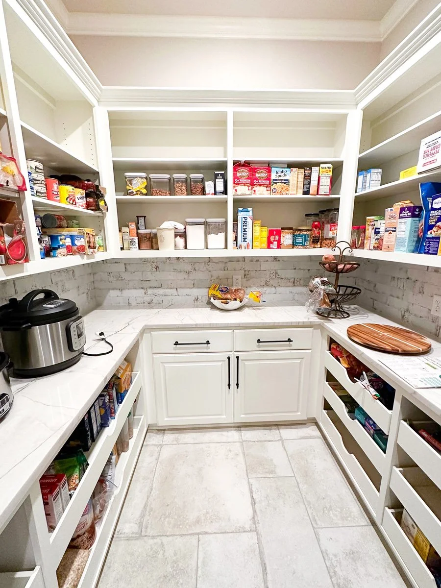 Pantry with white shelving units filled with food containers, boxes, and jars, a white countertop with a covered dish and a wooden cutting board, a black pressure cooker, and a white cabinet below the counter, tiled floor.