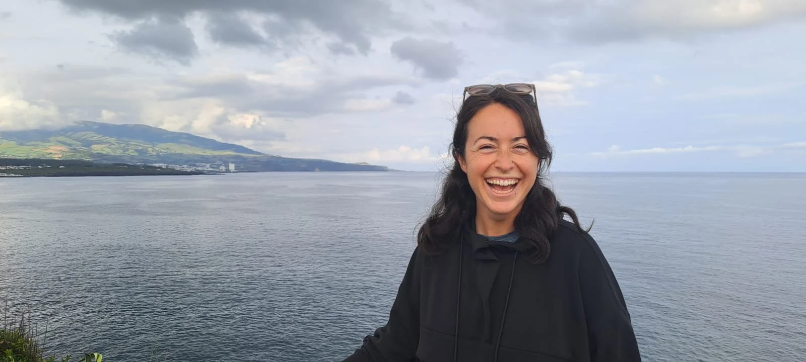 Woman with sunglasses smiling in front of a body of water and distant mountains under a cloudy sky.