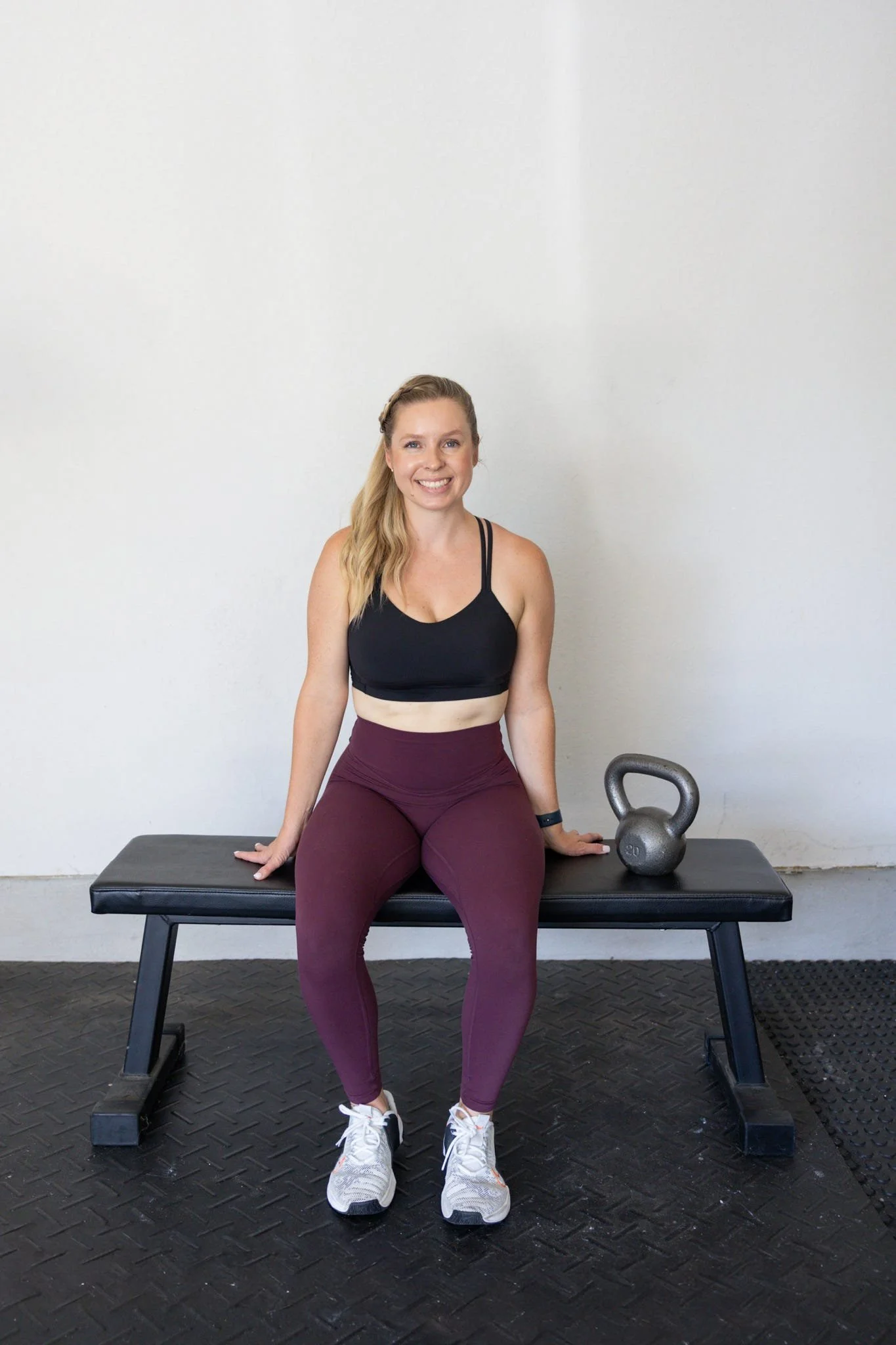 A woman with long blonde hair sitting on a black workout bench, smiling. She is dressed in a black sports bra, maroon leggings, and white athletic shoes. There is a kettlebell and a gray water bottle on the bench to her right. The background features a white wall and a black rubber gym floor.