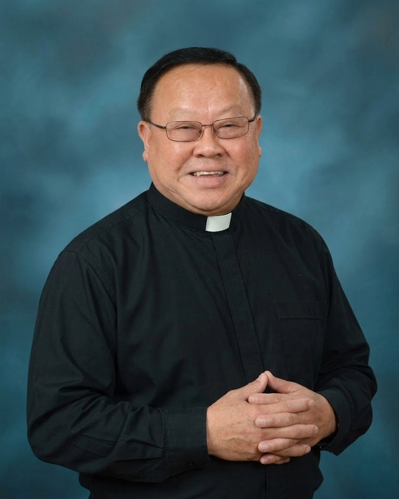 A smiling priest wearing glasses and a black clerical shirt with a white collar, standing against a blue background.
