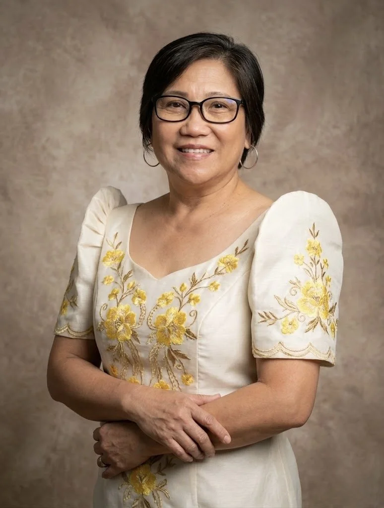 A middle-aged woman with short black hair, glasses, and hoop earrings, smiling and standing with her arms crossed in front of a neutral background. She is wearing a cream-colored dress with yellow floral embroidery.