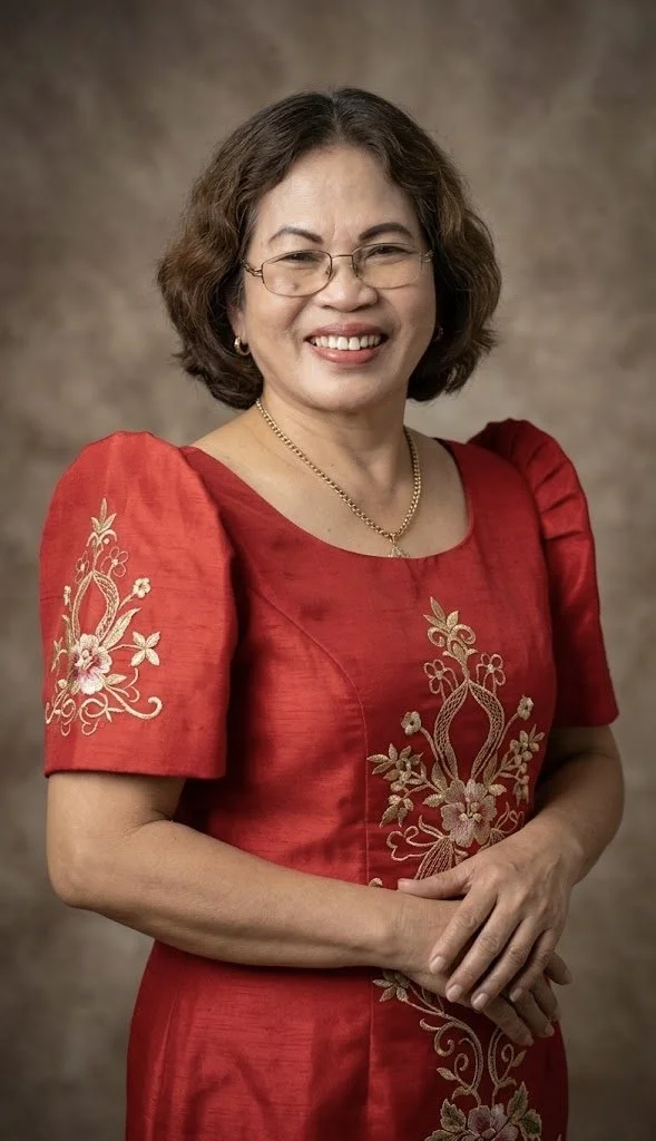A smiling woman with short curly hair and glasses, wearing a red traditional dress with floral embroidery, standing against a neutral background.