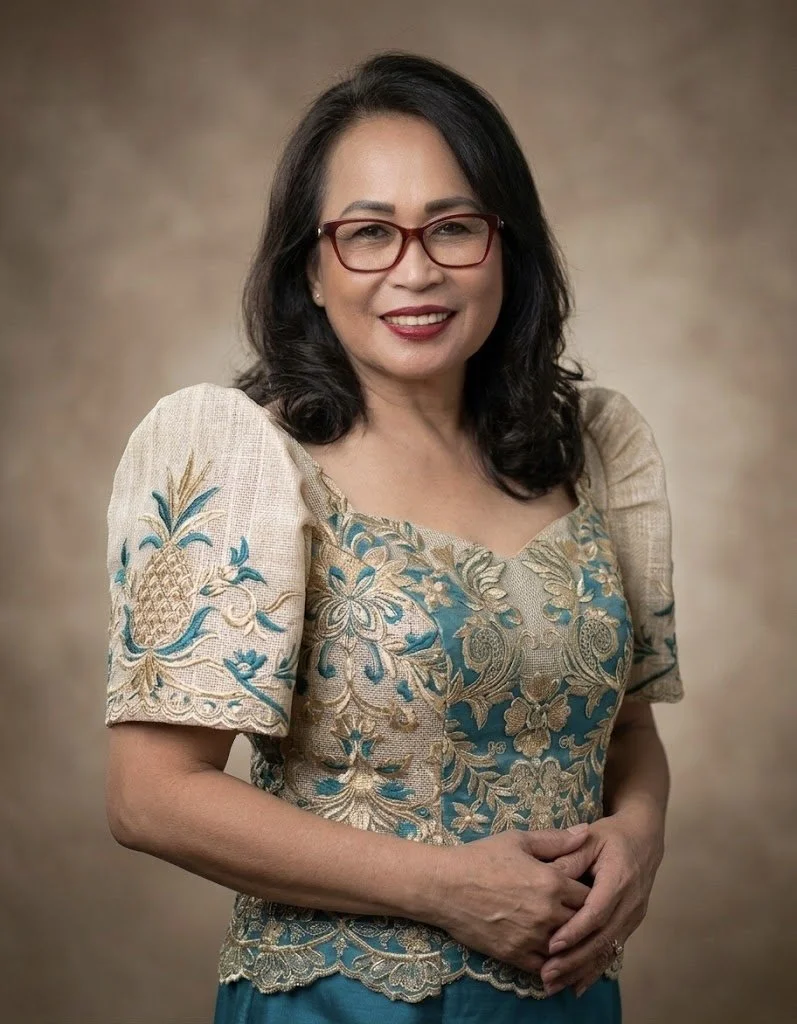 A woman with black hair, glasses, and red lipstick smiling, wearing a beige and teal embroidered dress with floral and pineapple motifs, standing against a brownish background.