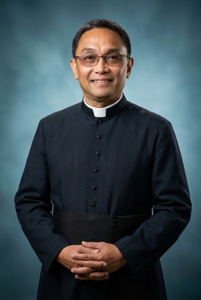 A man wearing a black clergy cassock with a white clerical collar, glasses, and short black hair, standing against a blue gradient background and smiling.