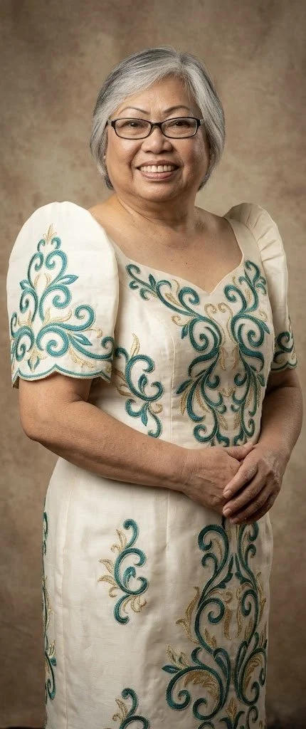 An older woman with short gray hair, wearing glasses and a white dress with blue and gold embroidery, standing against a brown textured background.