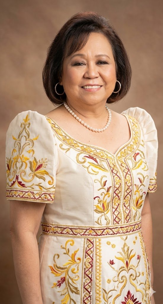 A woman with short dark hair, wearing hoop earrings, a pearl necklace, and a white dress with yellow, red, and gold embroidery, standing against a brown background.