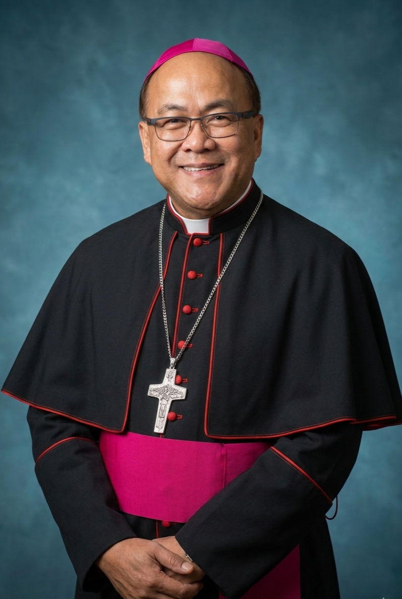 A smiling man dressed in Catholic bishop attire, wearing a black cassock with red piping, a pink sash, a large silver cross necklace, glasses, and a pink zucchetto, standing against a blue background.