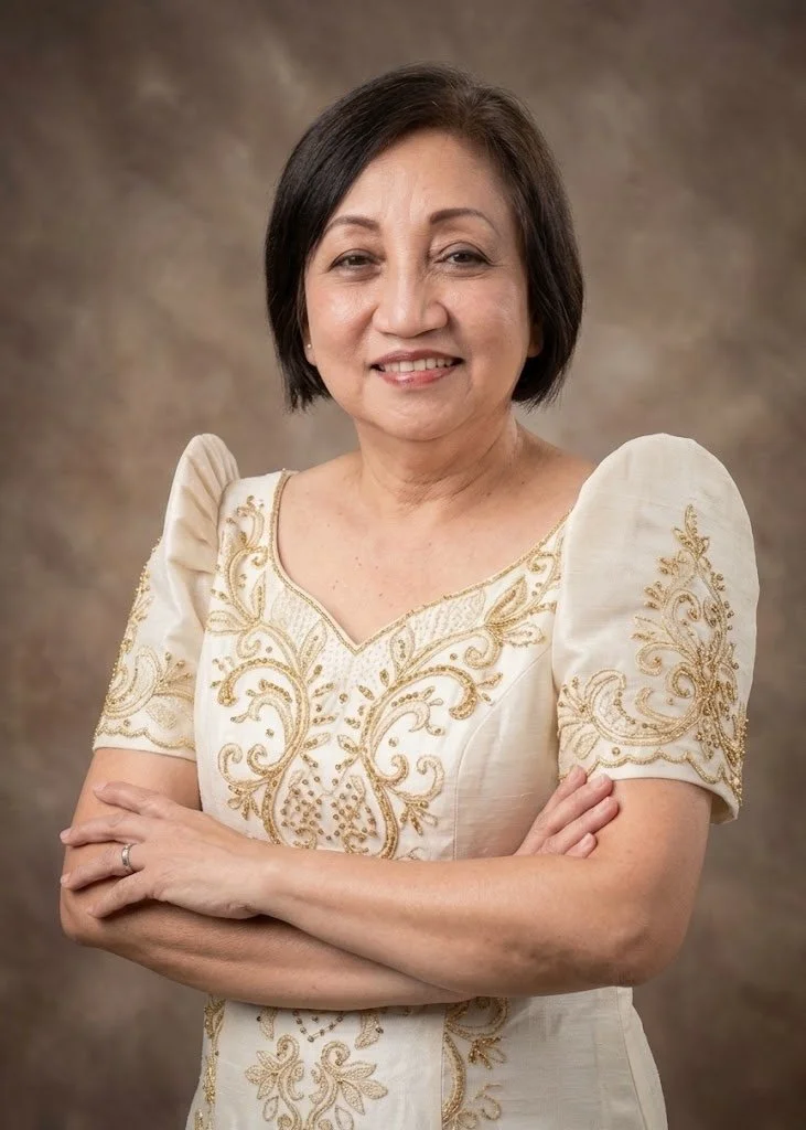 A middle-aged woman with short dark hair, smiling, wearing a cream-colored dress with gold embroidery, standing with her arms crossed against a brown background.