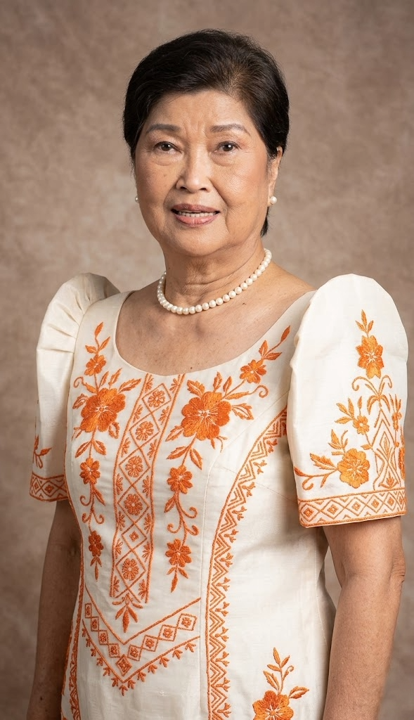 A woman with short black hair wearing a white dress with orange floral embroidery, a pearl necklace, and pearl earrings, standing against a beige background.