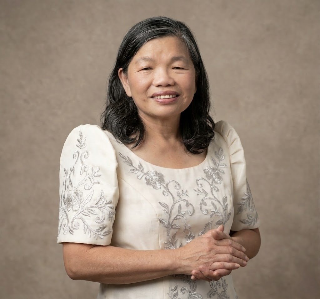 A middle-aged Asian woman with black hair and a gentle smile, standing with her hands clasped in front of her, wearing an ivory dress with embroidered silver floral patterns, against a neutral background.
