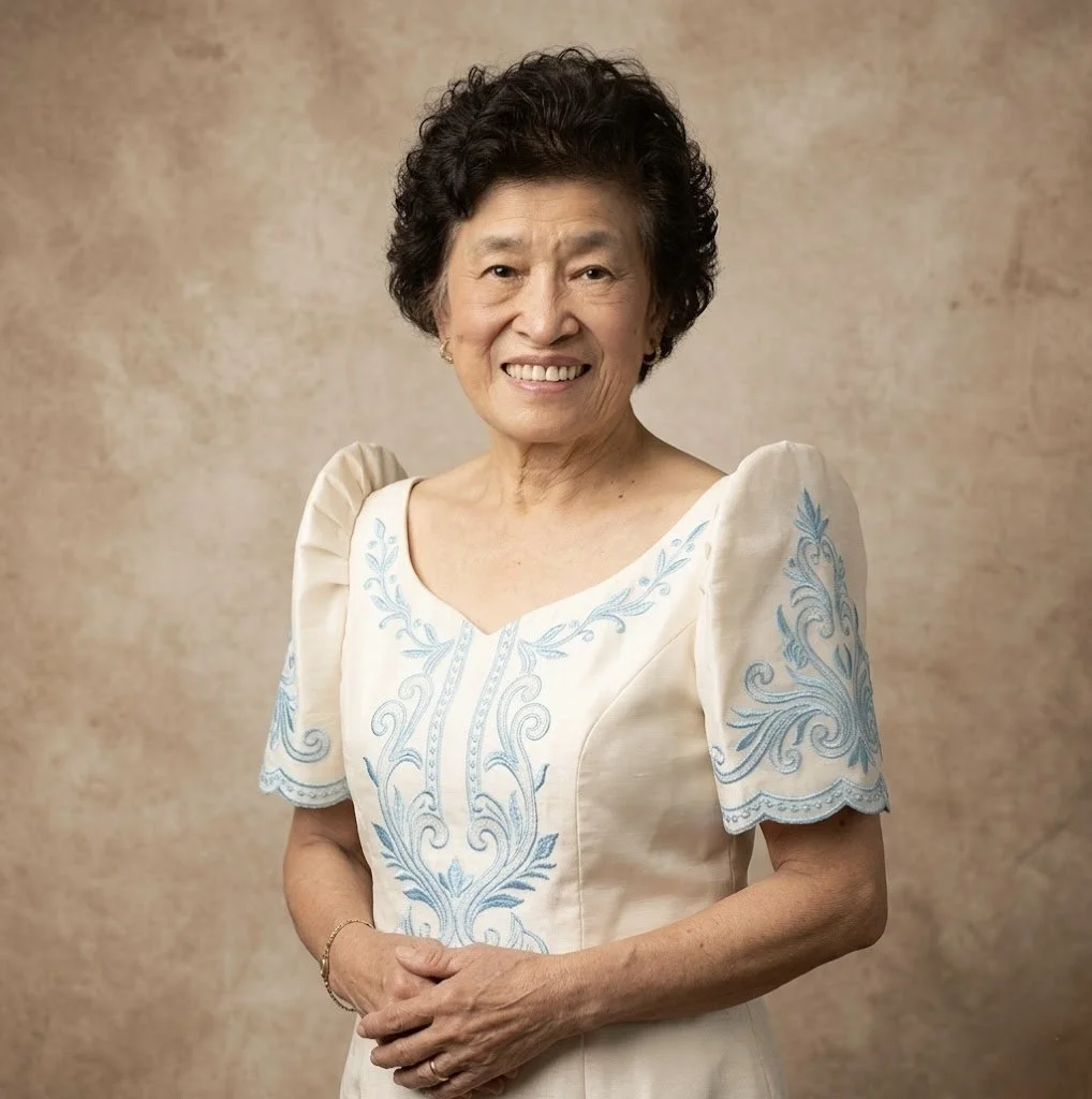 An elderly woman with short, curly black hair, smiling, wearing a cream-colored dress with light blue embroidery, standing against a beige background.