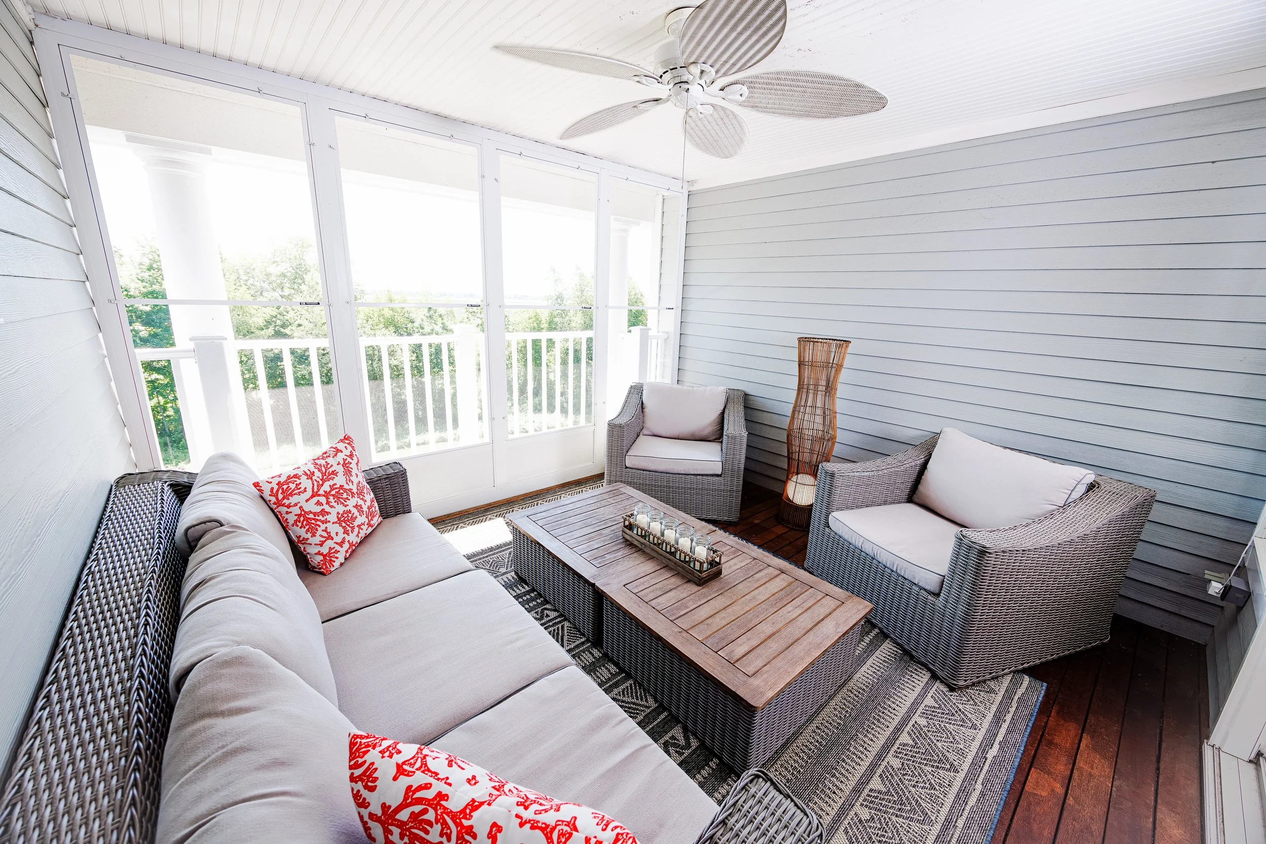 Sunlit screened porch with wicker furniture, including a sofa with red coral-patterned pillows, two armchairs, a wooden coffee table, a woven floor lamp, and a ceiling fan, overlooking a green landscape.