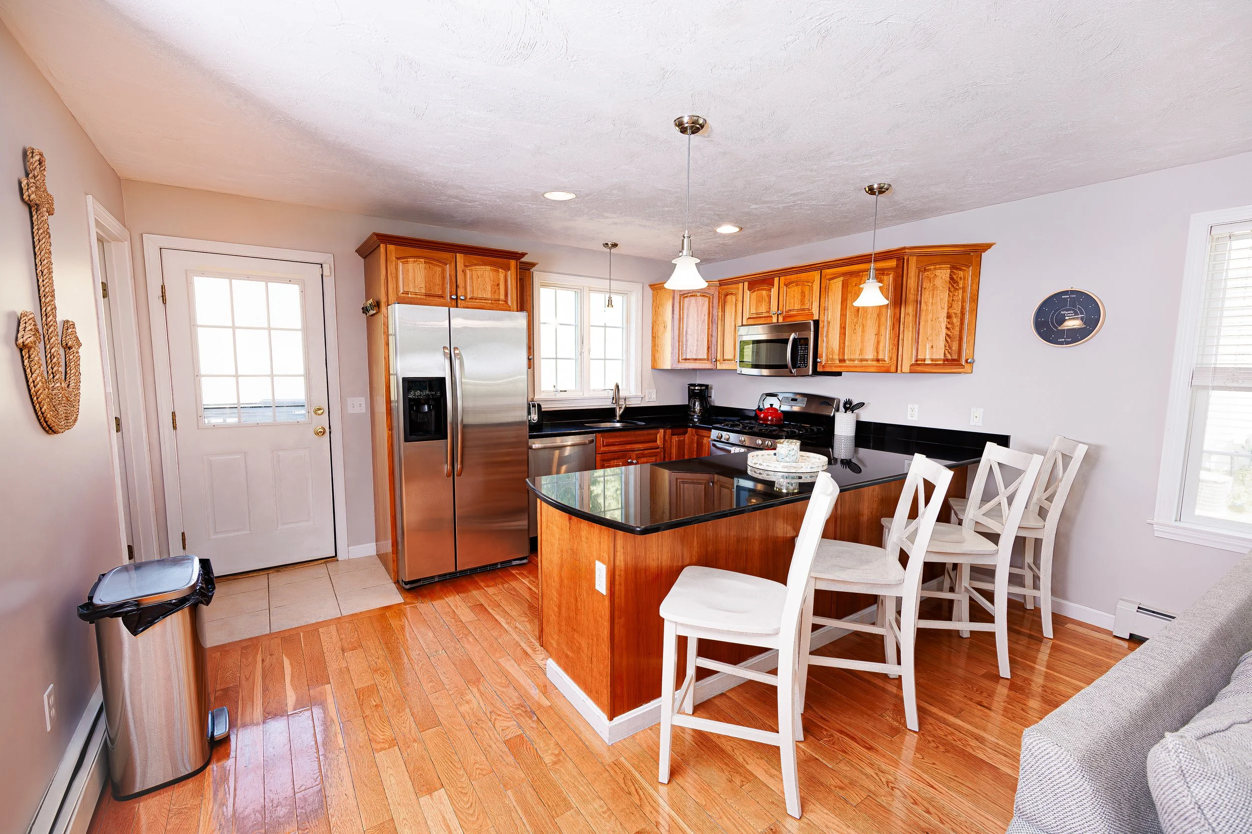 Kitchen with wooden cabinets, black countertops, stainless steel refrigerator, microwave, gas stove, small kitchen island, white bar stools, and a couch partially visible on the right.