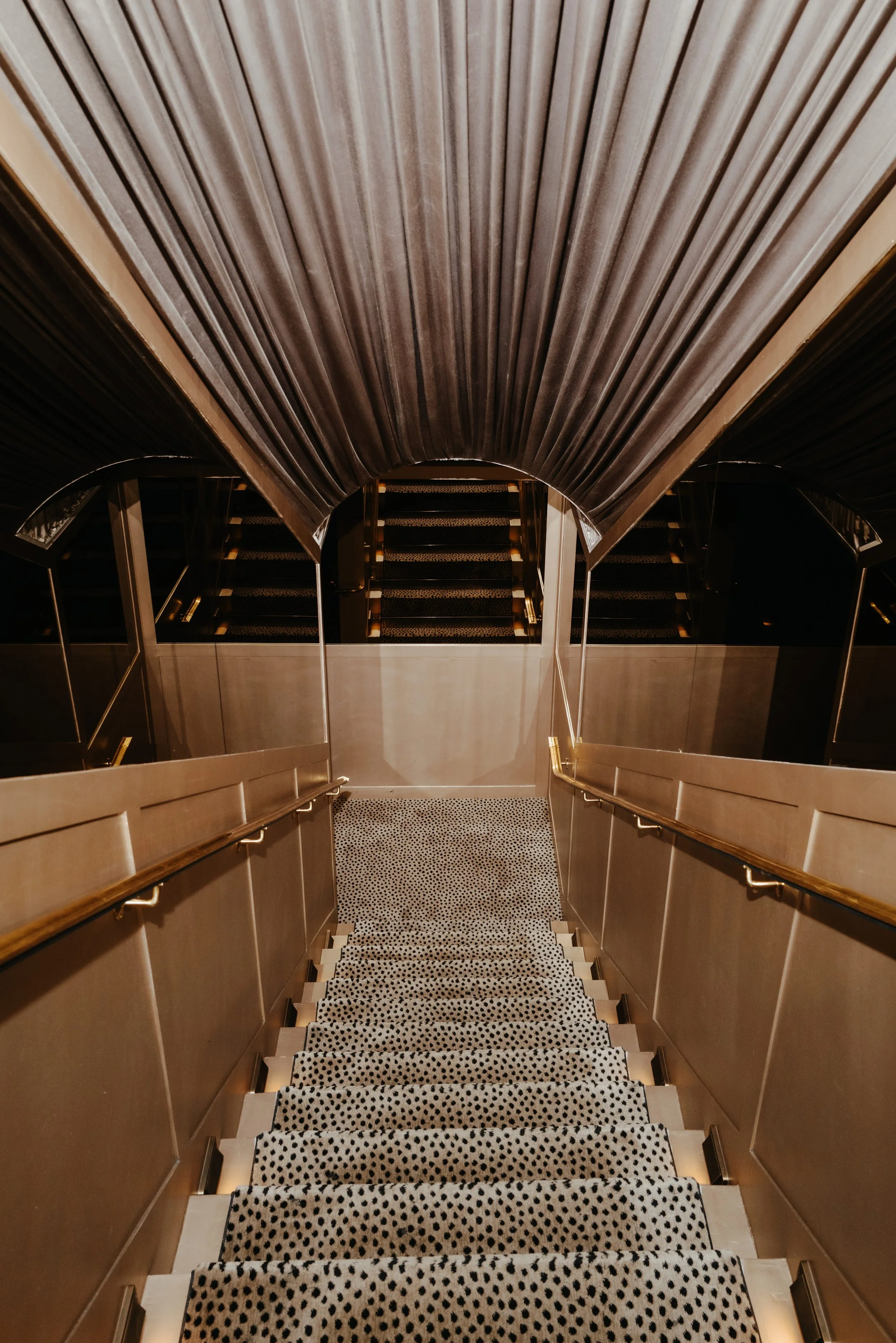 View of a carpeted staircase with black and white polka dot pattern, flanked by beige walls with wooden handrails on both sides, leading up to a landing with closed curtains covering a window or wall.