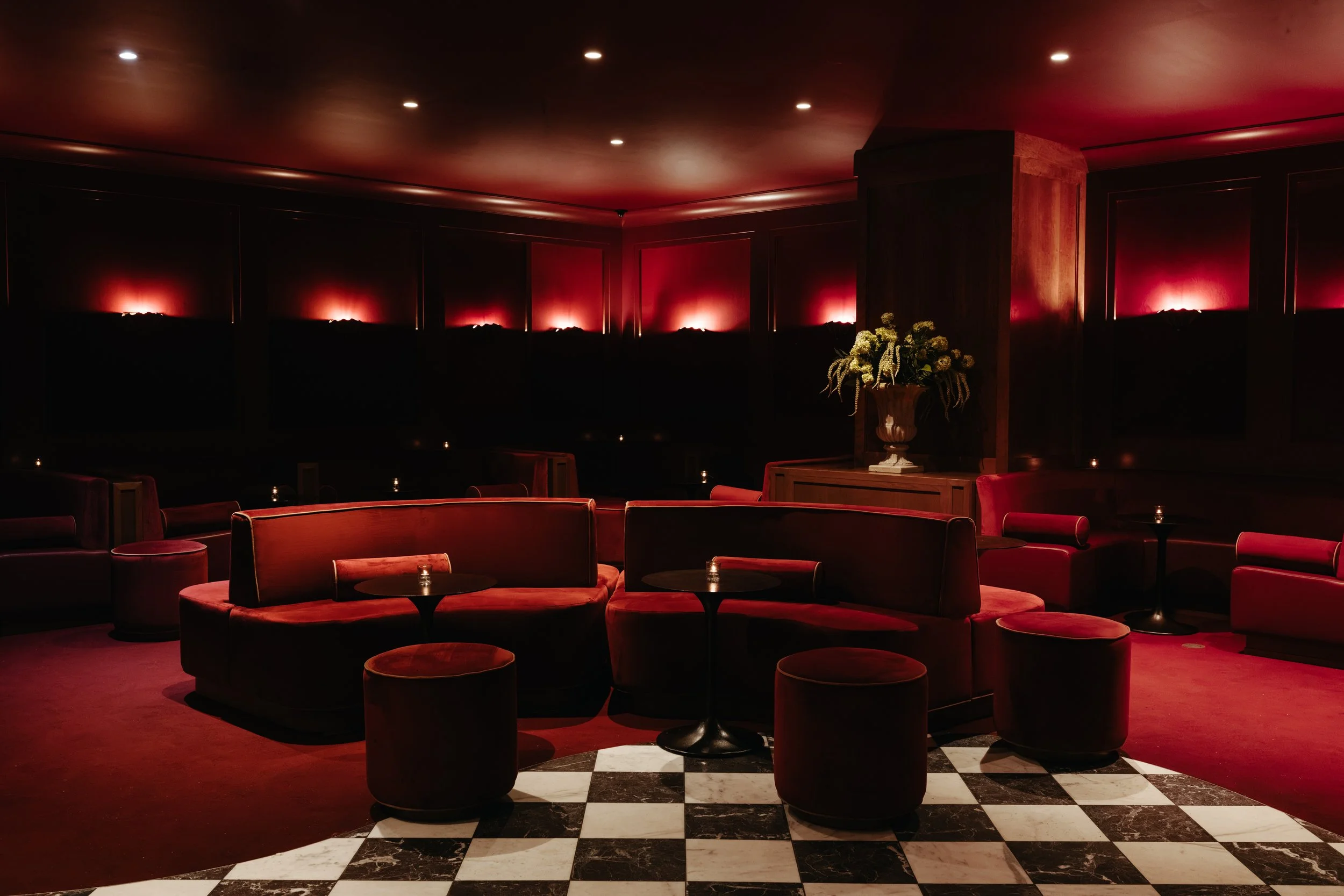 A dimly lit lounge with red chairs, small round tables, a black and white checkered floor, a large flower arrangement, and red-lit wall panels. BC Lounge Club