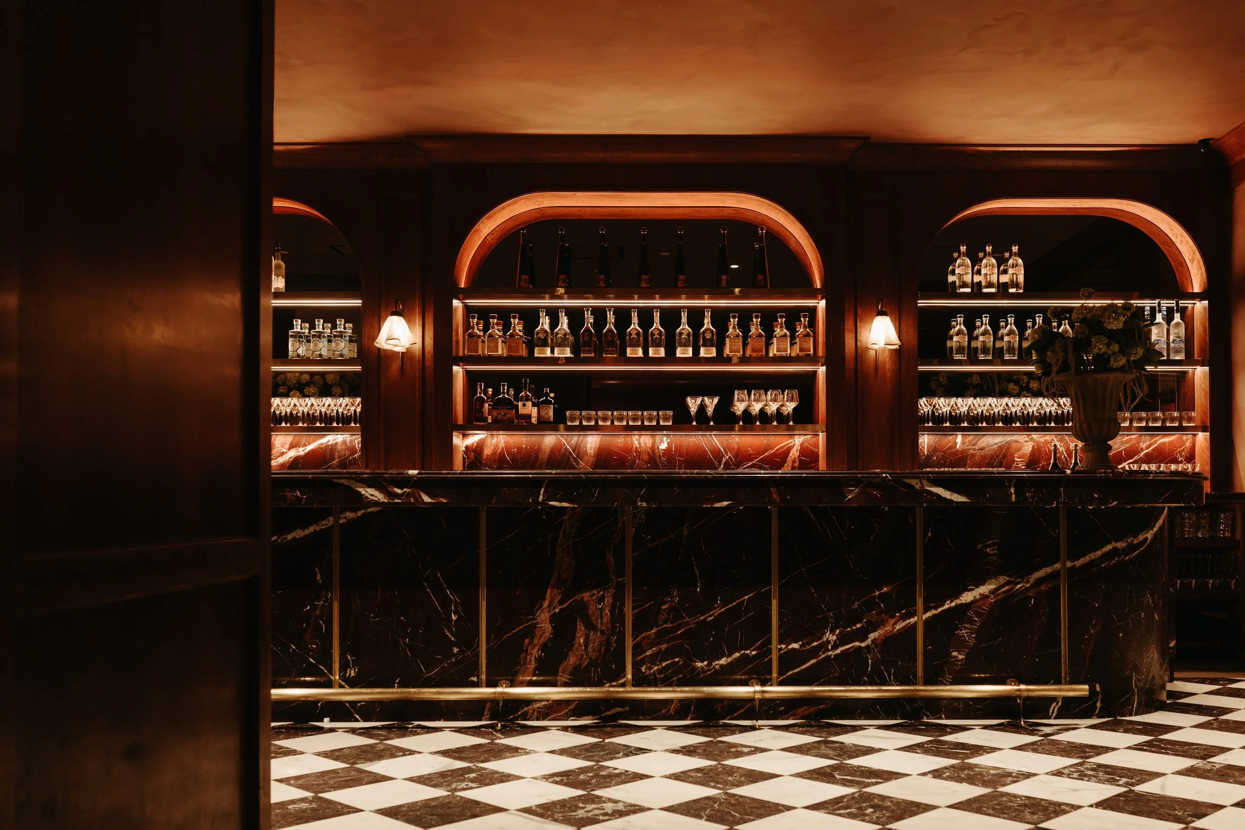 Elegant bar with dark wood shelves displaying bottles of liquor, illuminated with warm lighting, and a vase with flowers on a marble counter with a black and white checkered floor. BC Lounge Club