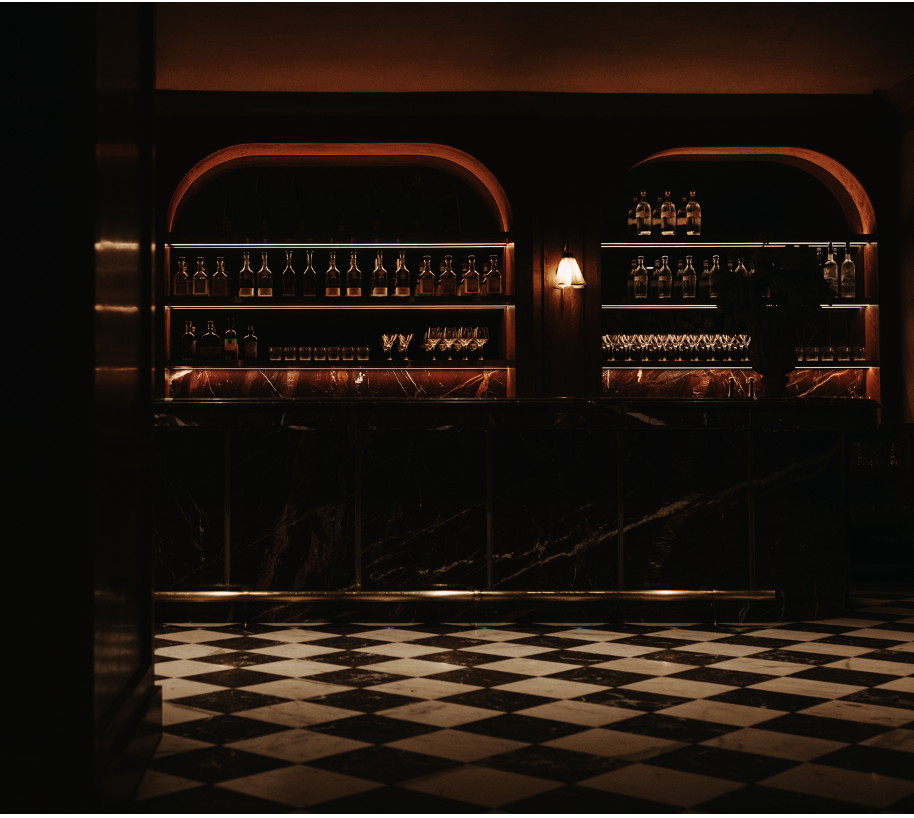 Dimly lit bar with shelves of liquor bottles and glassware, dark marble counter, checkered black and white floor. BC Club & Lounge