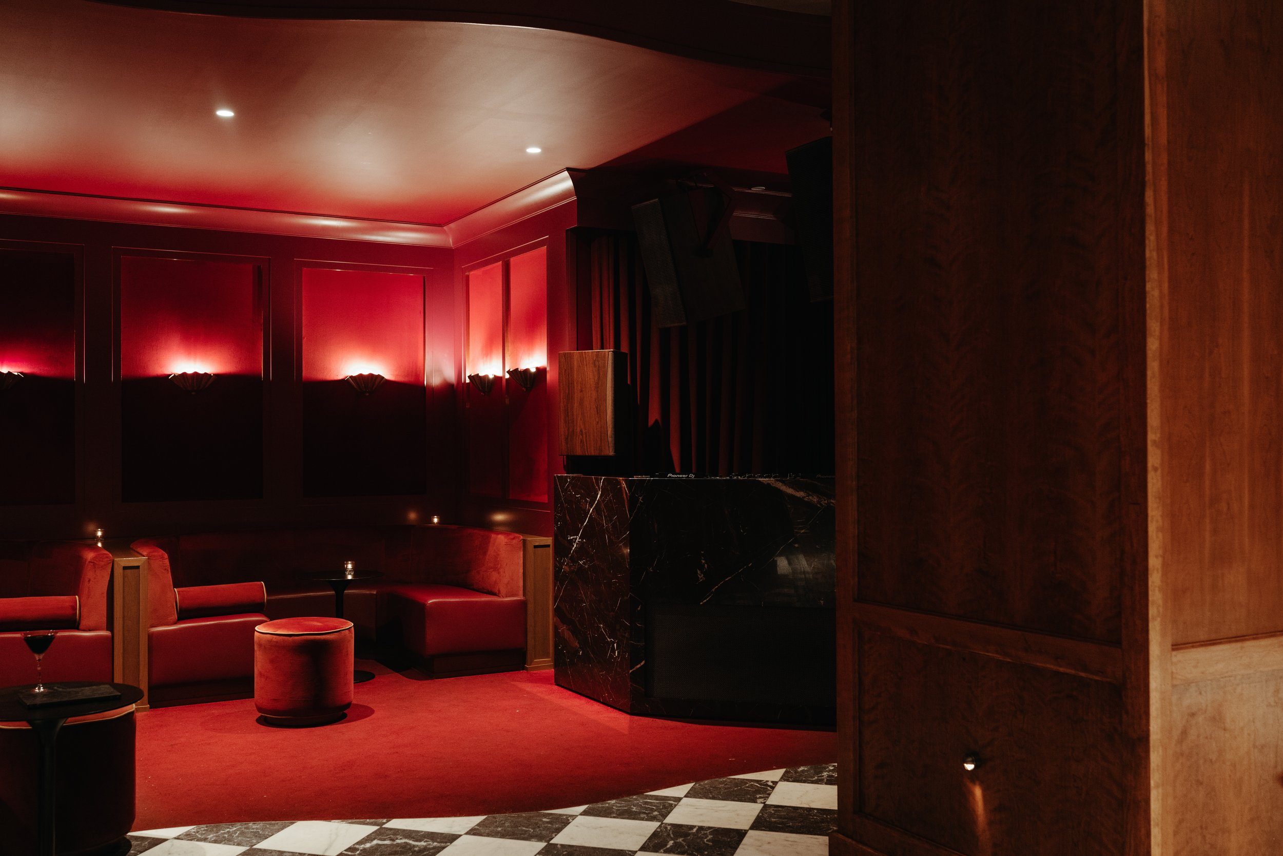 Interior of a lounge or bar area with red upholstered chairs, black marble tables, red wall with sconces, and dark wood paneling. BC Lounge Club