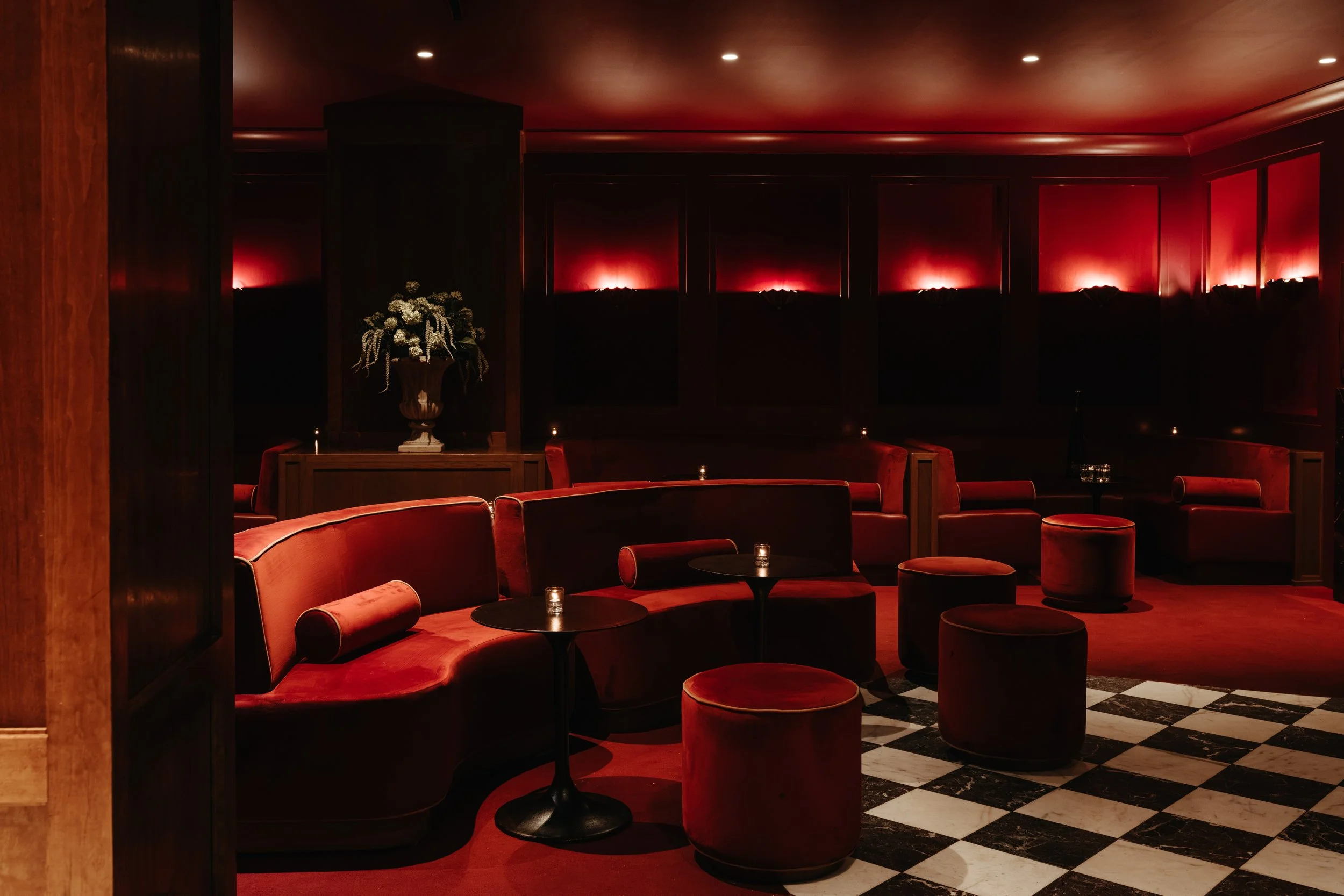 A dimly lit lounge area with red velvet chairs, small round tables, and a black-and-white checkered floor. A large floral arrangement is on a table, and red lighting highlights the dark wood-paneled wall. BC Lounge Club