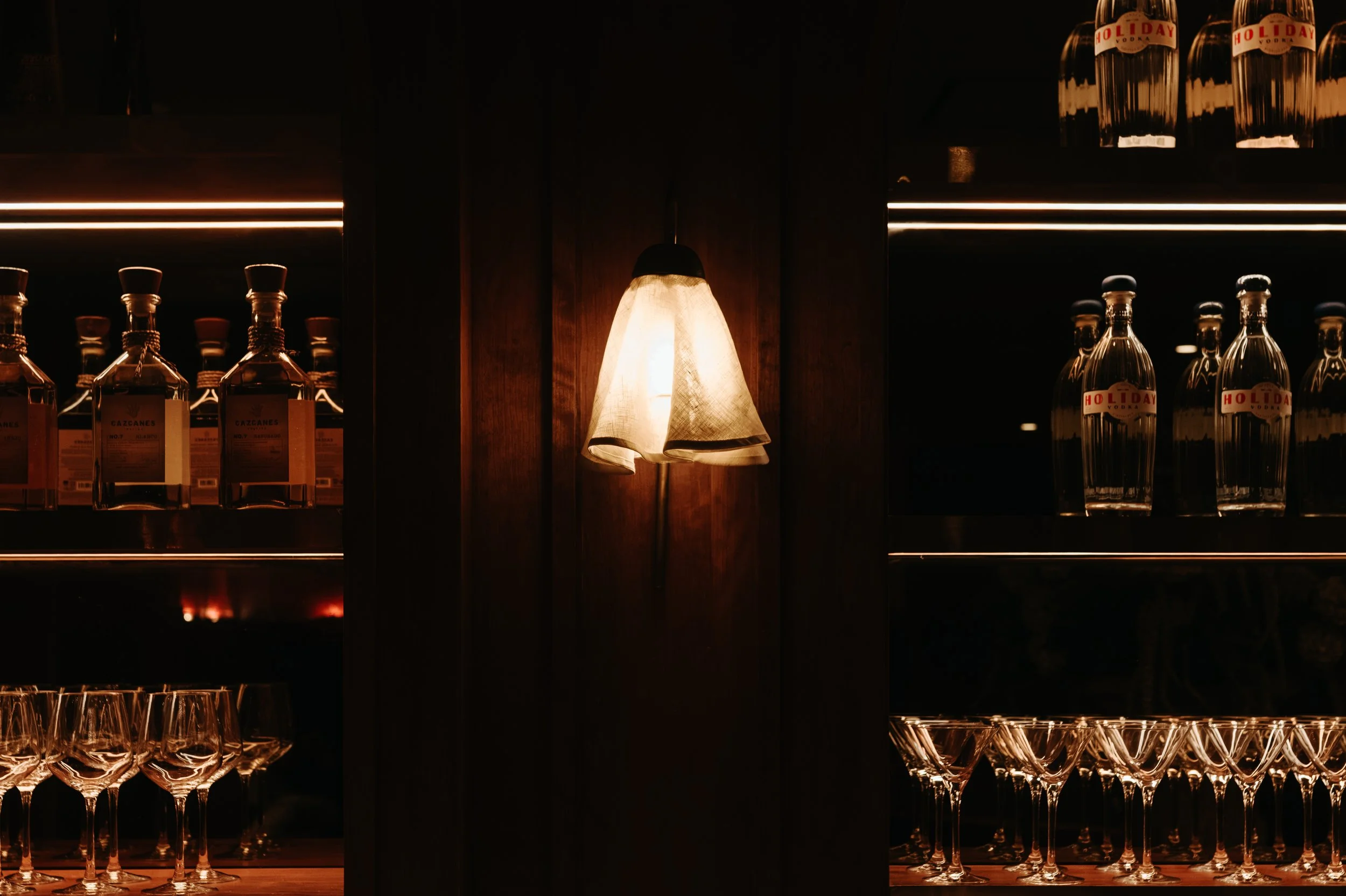 Dark bar with shelves of liquor bottles, pendant light in the center, glassware at the bottom, and illuminated bottles on the shelves.