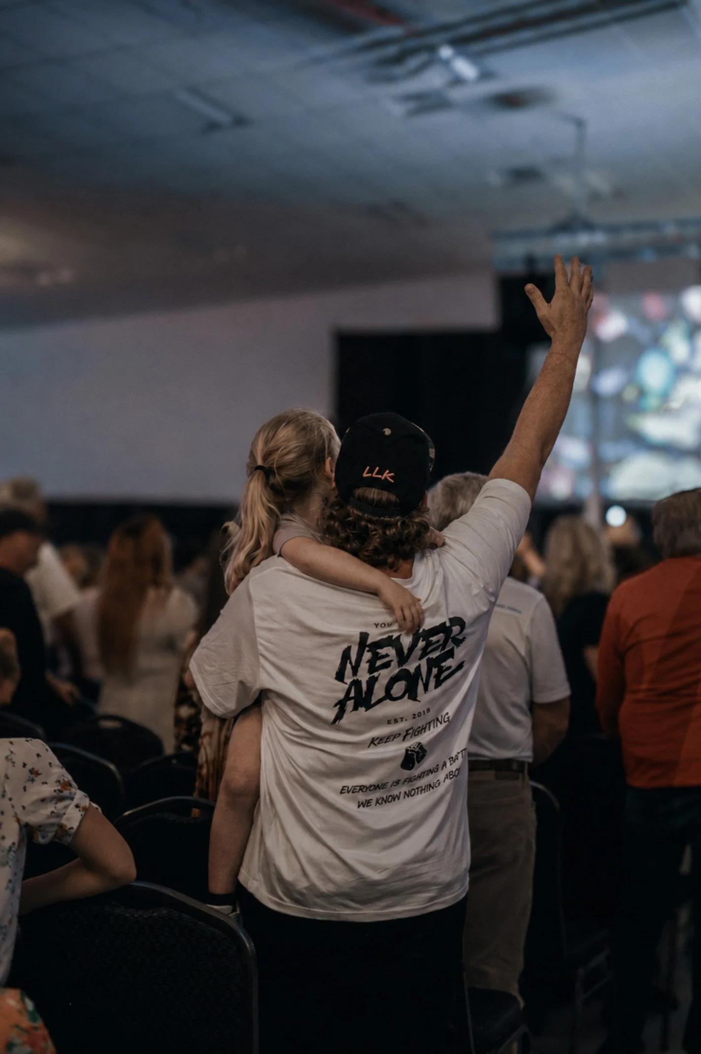A group of people gathered indoors, with a person in the center holding a small child while raising their hand. The group appears to be attending an event or rally. The person holding the child is wearing a white T-shirt with text on the back that reads "NEVER ALONE," and the child has blonde hair tied in a ponytail.