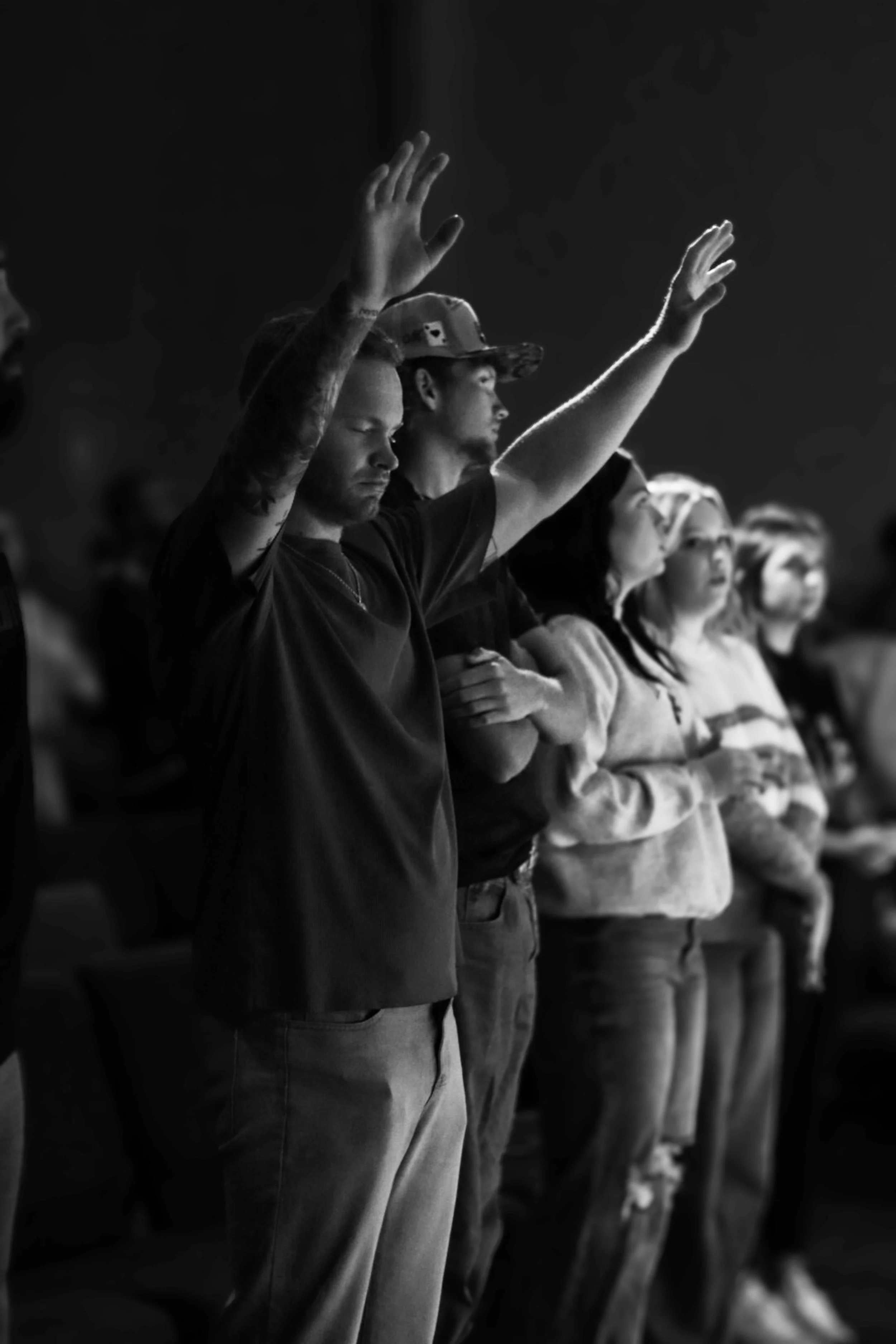People in a line with eyes closed, some raising their hands, in a dark room, participating in a spiritual or meditative event.