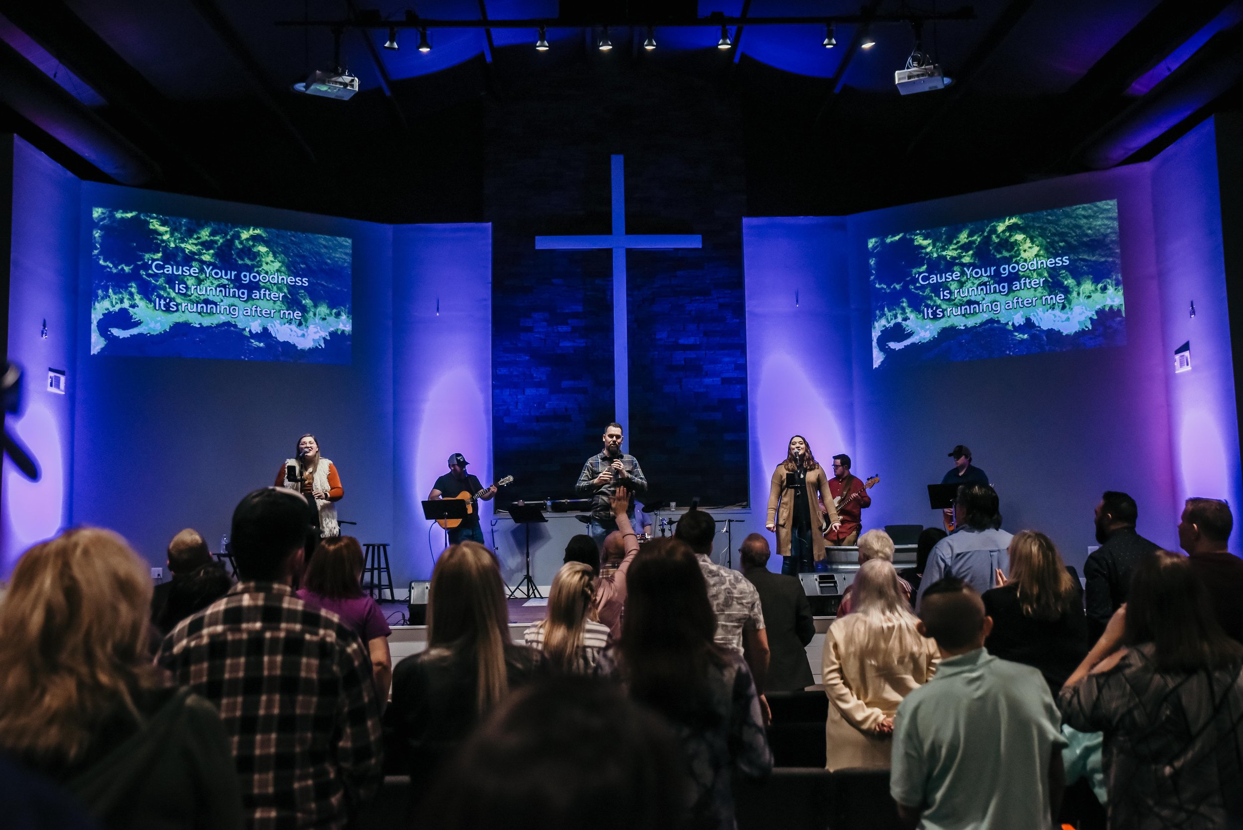 A church service or concert with a group of people singing and playing instruments on stage. There's a large cross and two screens displaying lyrics, with audience members in the foreground.