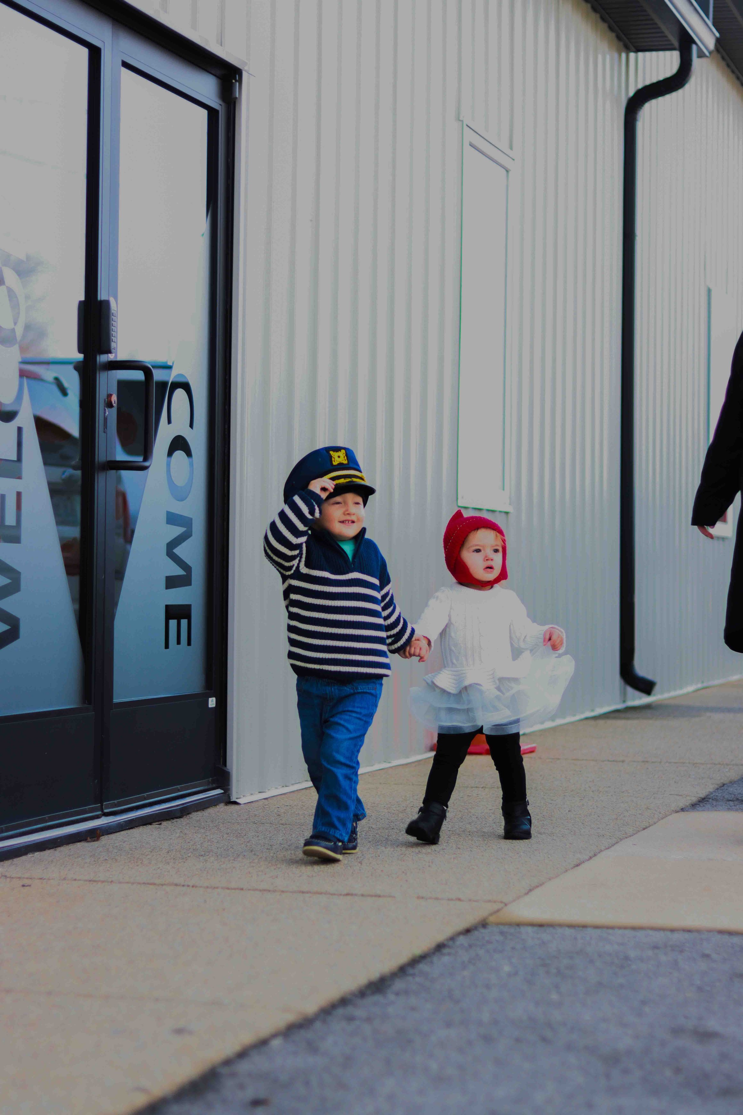 Two young children holding hands, walking outside near a building with metal siding. The boy is wearing a police hat and a striped sweater, and the girl is wearing a red hat and white dress.