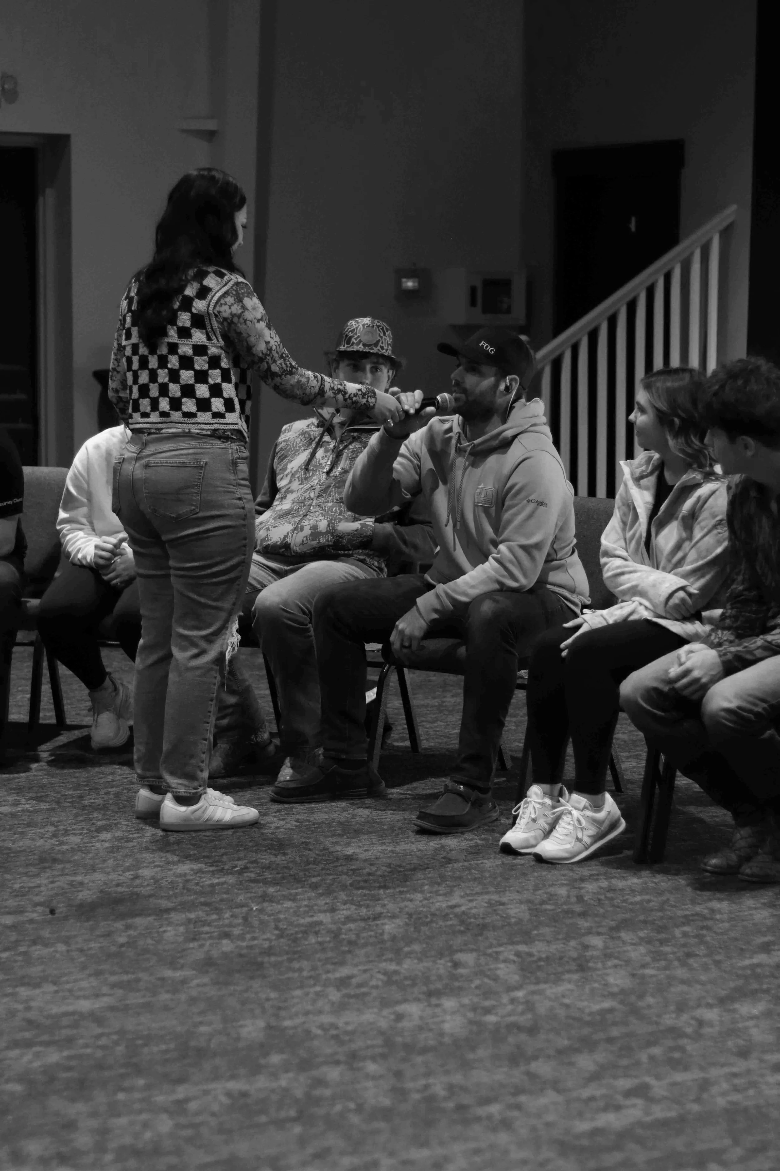 A woman is holding a microphone to a man who is speaking during a group interview or discussion. Several people are seated around them, with some looking at the man and others looking elsewhere, in a casual indoor setting.